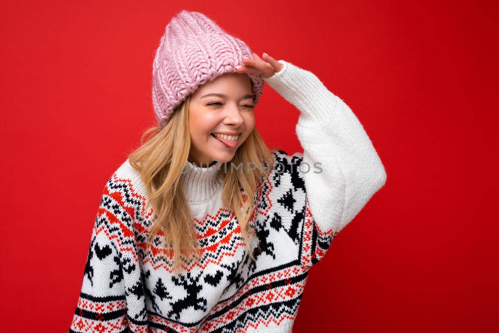 Photo of young positive happy joyful attractive blonde woman with sincere emotions wearing knitted hat and warm winter jersey isolated over red background with empty space and looking to the side with hand.
