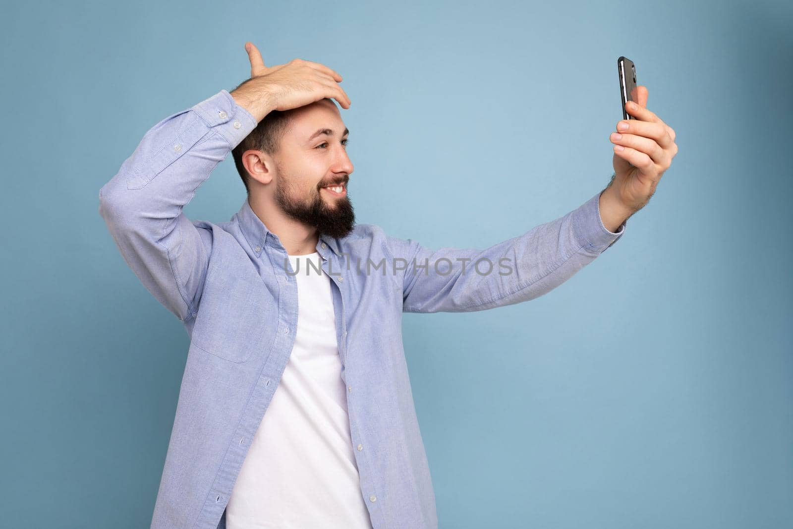Handsome young brunet bearded man wearing casual stylish clothes standing isolated over blue background wall holding smartphone taking selfie photo looking at mobile phone screen display by TRMK