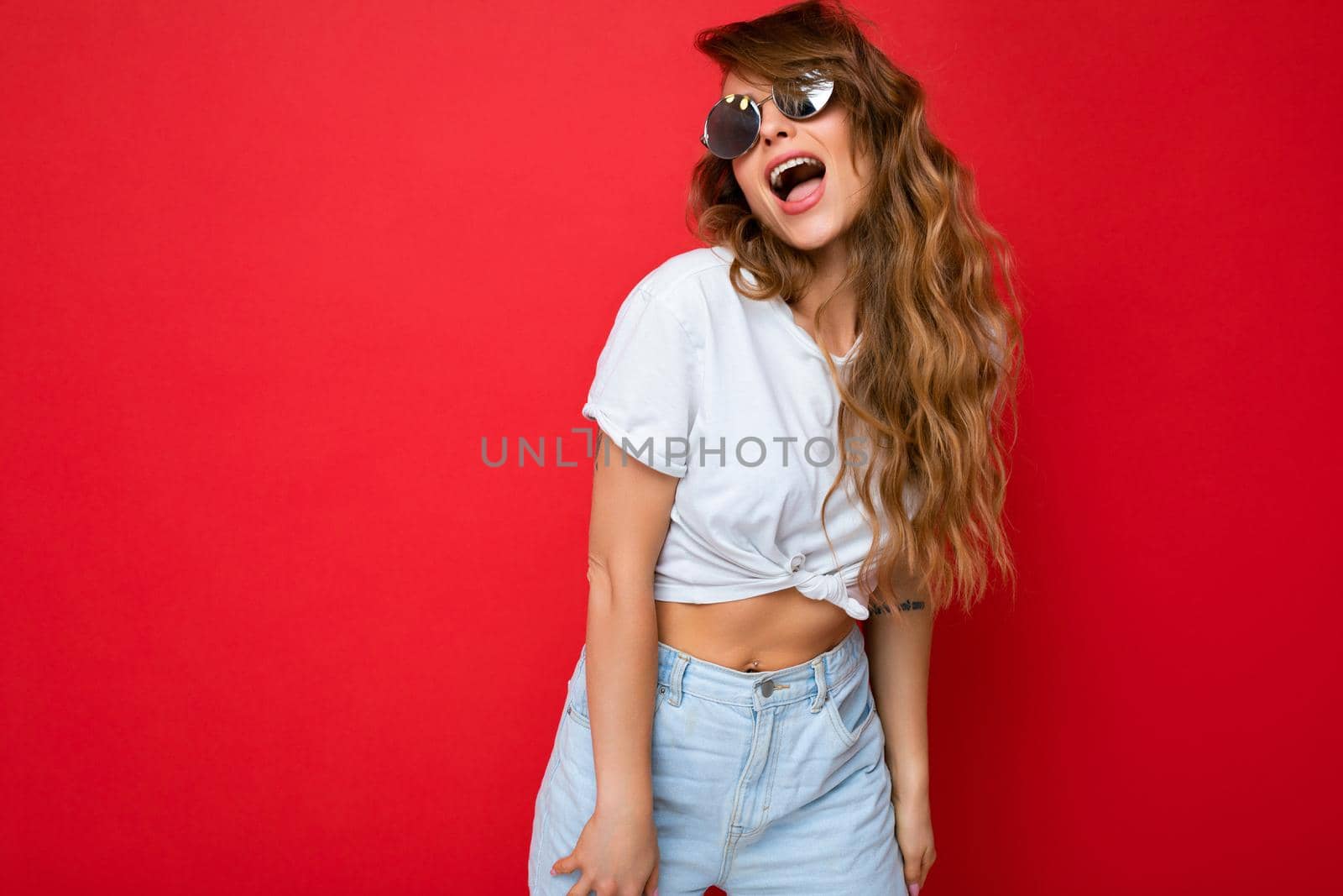 Shot of beautiful amazing happy young dark blonde curly woman isolated over red background wall wearing casual white t-shirt and stylish sunglasses looking to the side. copy space