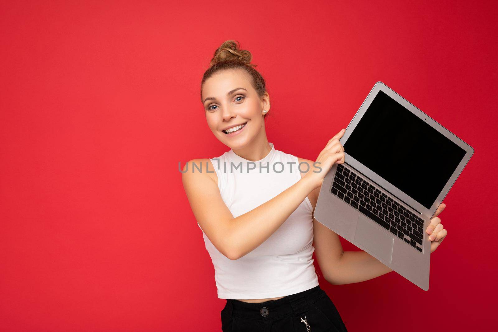 beautiful smiling fascinating happy blond young woman with gathered hair looking at camera holding computer laptop wearing white t-shirt isolated over red wall background.