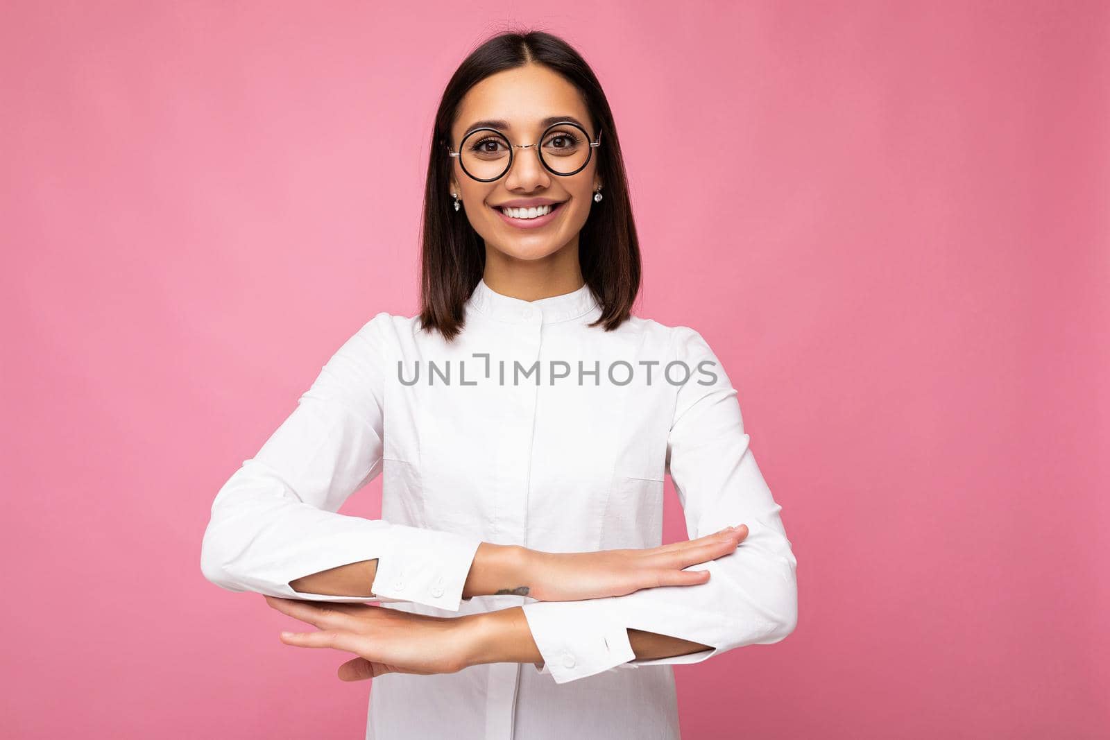Photo shot of beautiful young brunette woman wearing casual clothes and stylish optical glasses isolated over colorful background looking at camera imitating sitting at the desk.