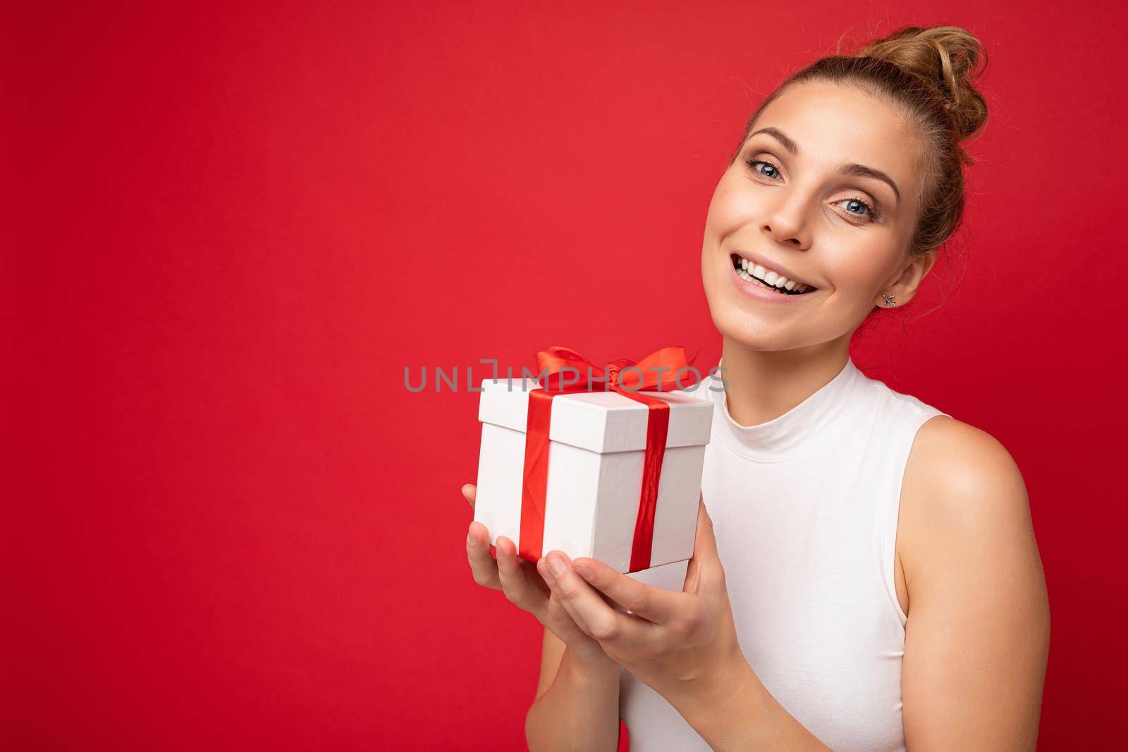 Beautiful happy young blonde woman isolated over colourful background wall wearing stylish casual clothes holding gift box and looking at camera by TRMK