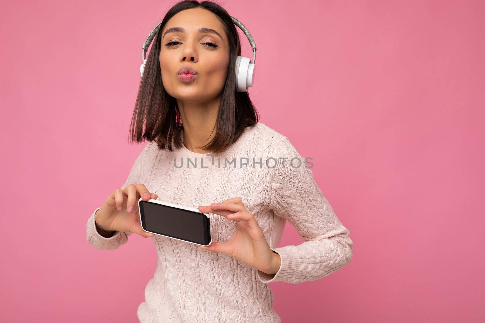 Attractive positive smiling young woman wearing stylish casual outfit isolated on colourful background wall holding and showing mobile phone with empty screen for cutout wearing white bluetooth headphones and having fun looking at camera and giving kiss.