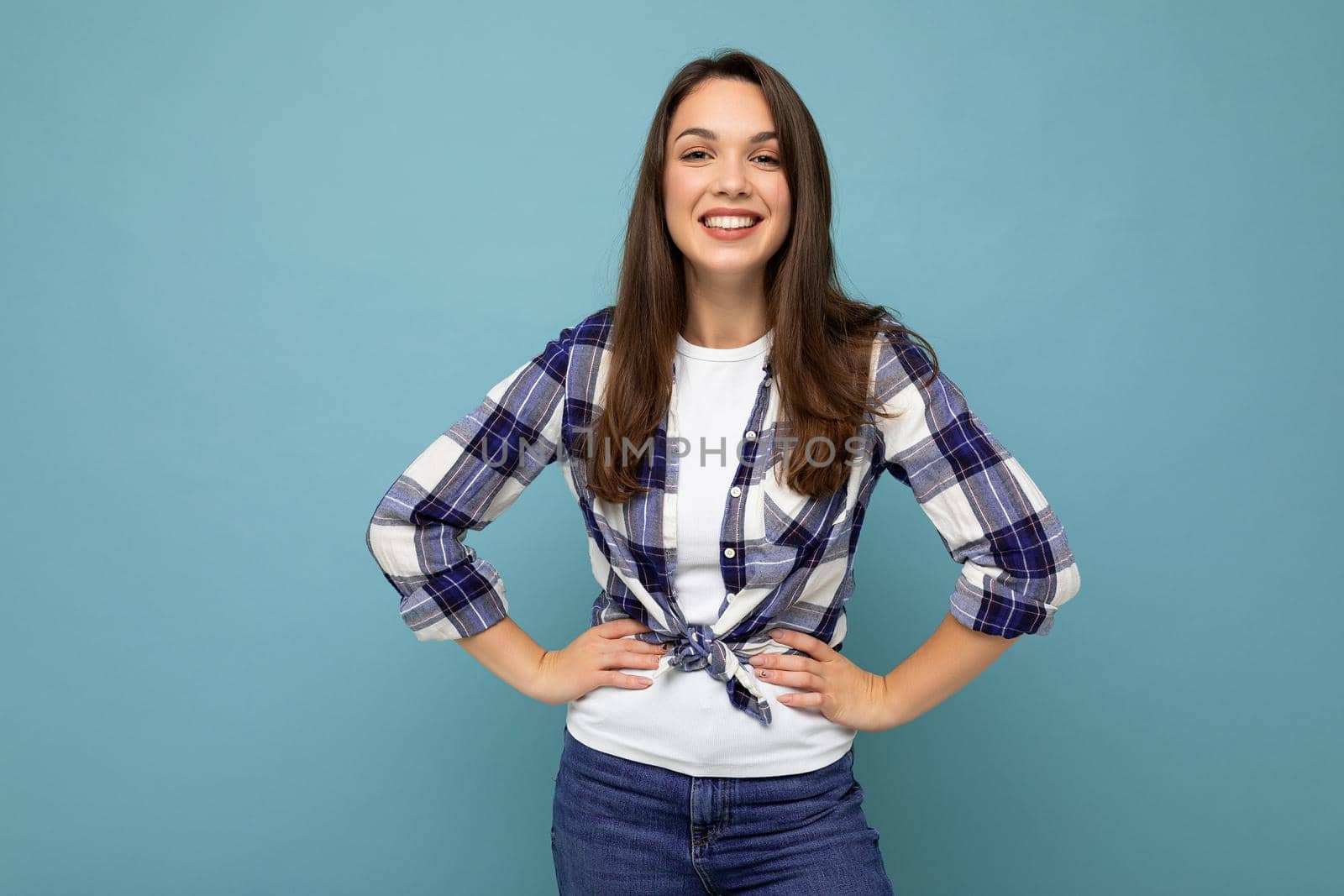 Photo portrait of young beautiful smiling hipster brunette woman in trendy blue and white shirt and jeans clothes. Sexy carefree female person posing isolated near blue wall with empty space in studio. Positive model with natural makeup.