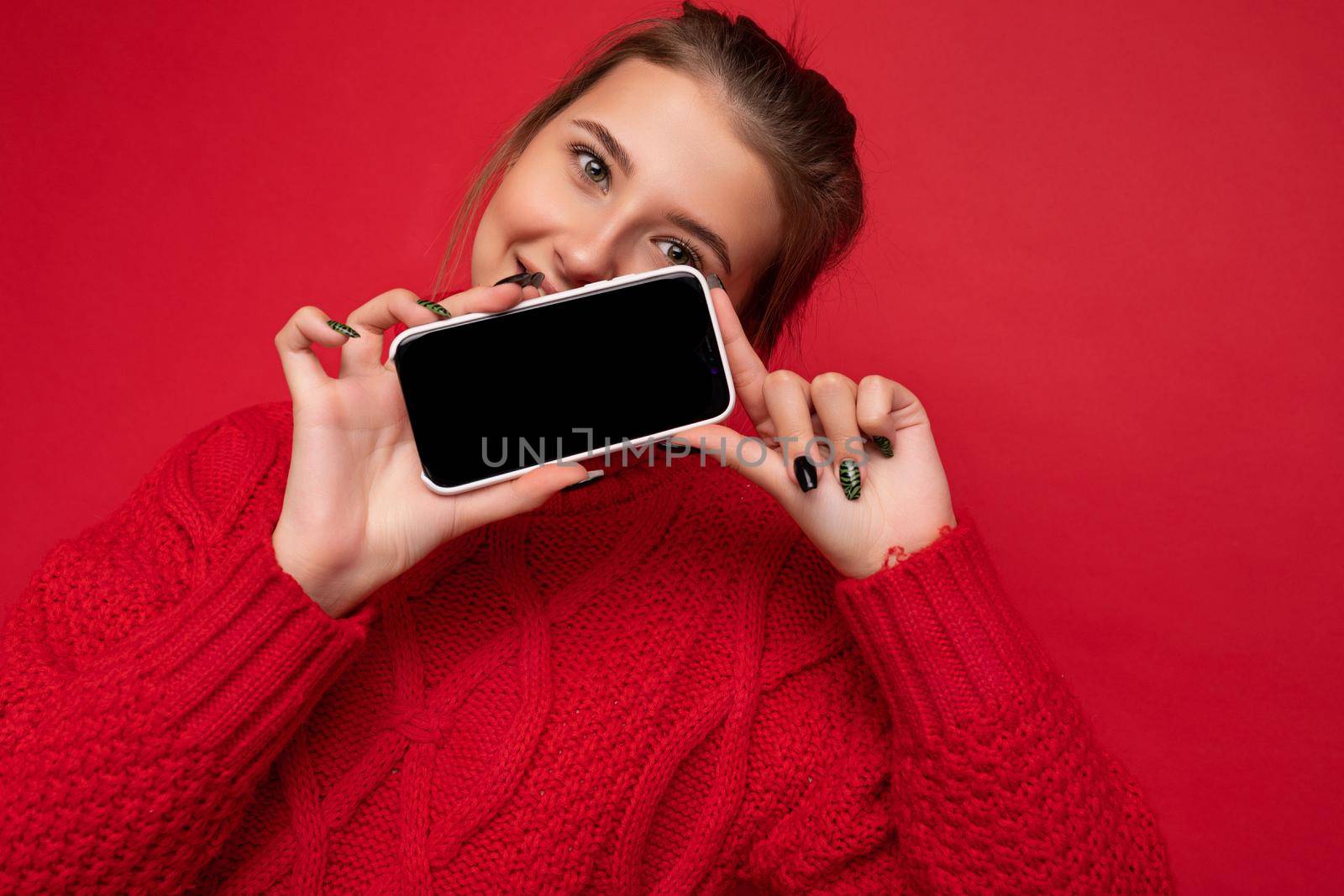 Photo of beautiful smiling young woman good looking wearing casual stylish outfit standing isolated on background with copy space holding smartphone showing phone in hand with empty screen display for mockup looking at camera.
