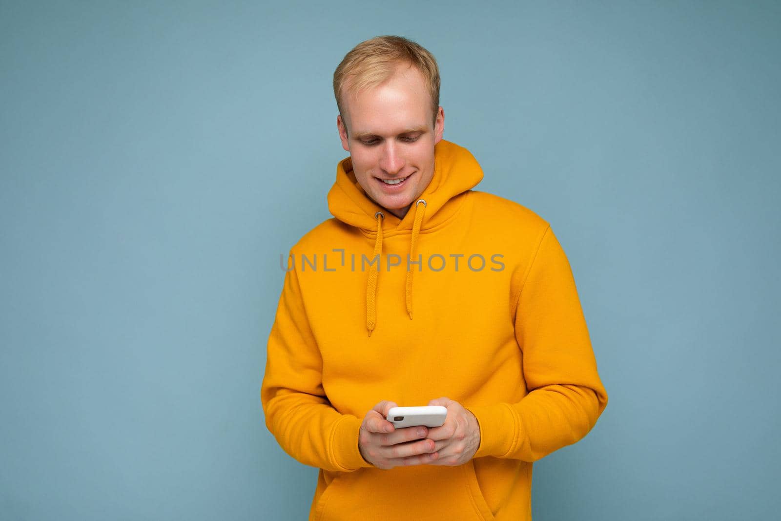 Photo shot of handsome positive good looking young man wearing casual stylish outfit poising isolated on background with empty space holding in hand and using mobile phone messaging sms looking at smartphone display screen by TRMK