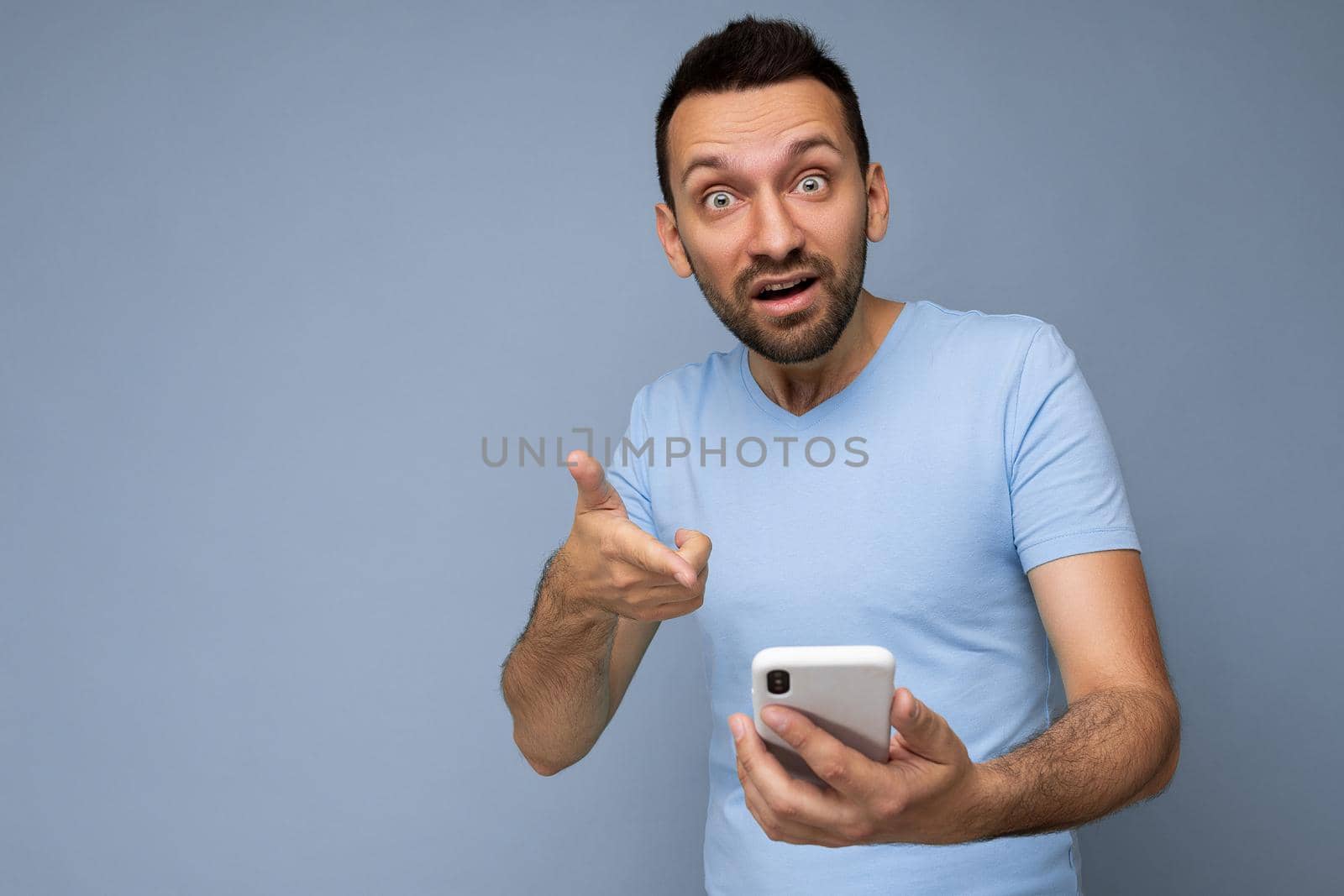 Photo of angry asking handsome young man with beard wearing everyday blue t-shirt isolated over blue background holding and using mobile phone communication online on the internet looking at camera.