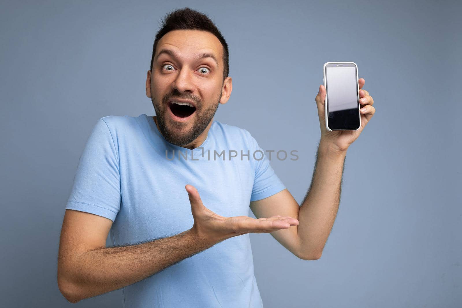 Amazed handsome young unshaven brunet man wearing everyday blue t-shirt isolated over blue background holding and showing mobile phone with empty display for cutout looking at camera and pointing at smartphone.
