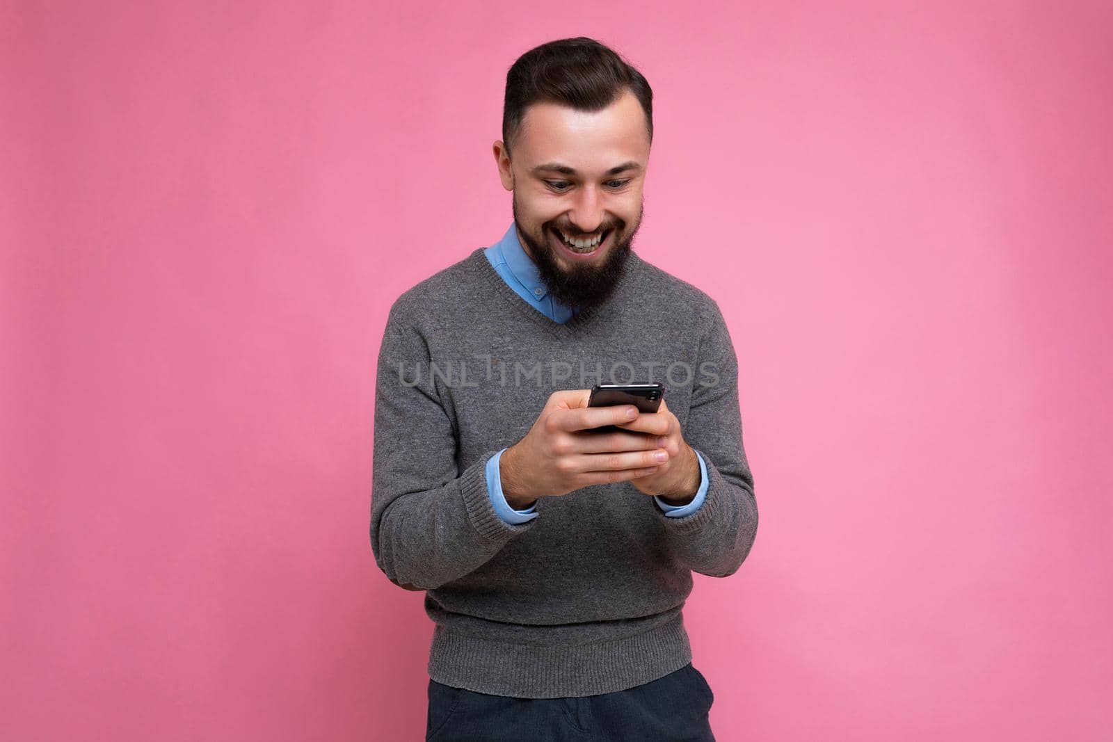 Positive smiling handsome good looking brunet bearded young man wearing grey sweater and blue shirt isolated on pink background with empty space holding in hand and using mobile phone communicating online looking at gadjet screen by TRMK