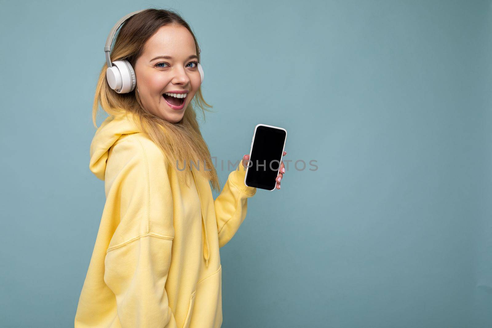 Side-profile photo of beautiful happy smiling young woman wearing stylish casual outfit isolated on background wall holding and showing mobile phone with empty display for mockup wearing white bluetooth headphones listening to music and having fun looking at camera and winking by TRMK