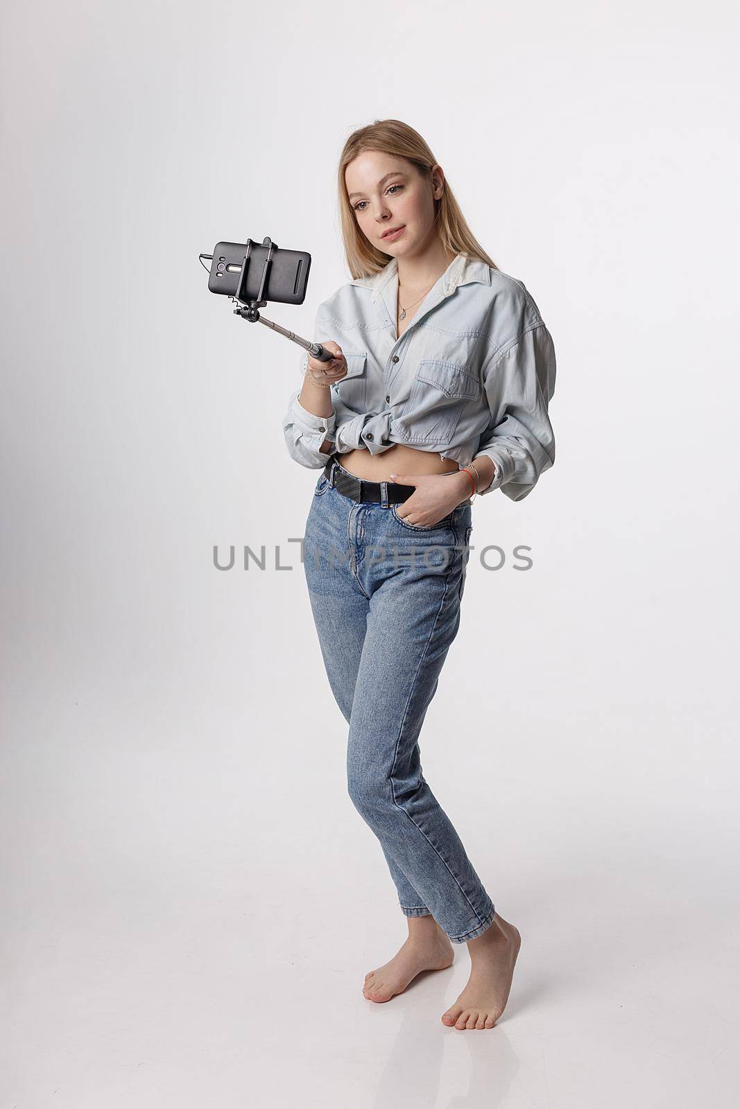 happy female making self portrait with smartphone attached to selfie stick pole. young pretty blond girl with long hair smiling and using cell phone isolated on white background. modern communication