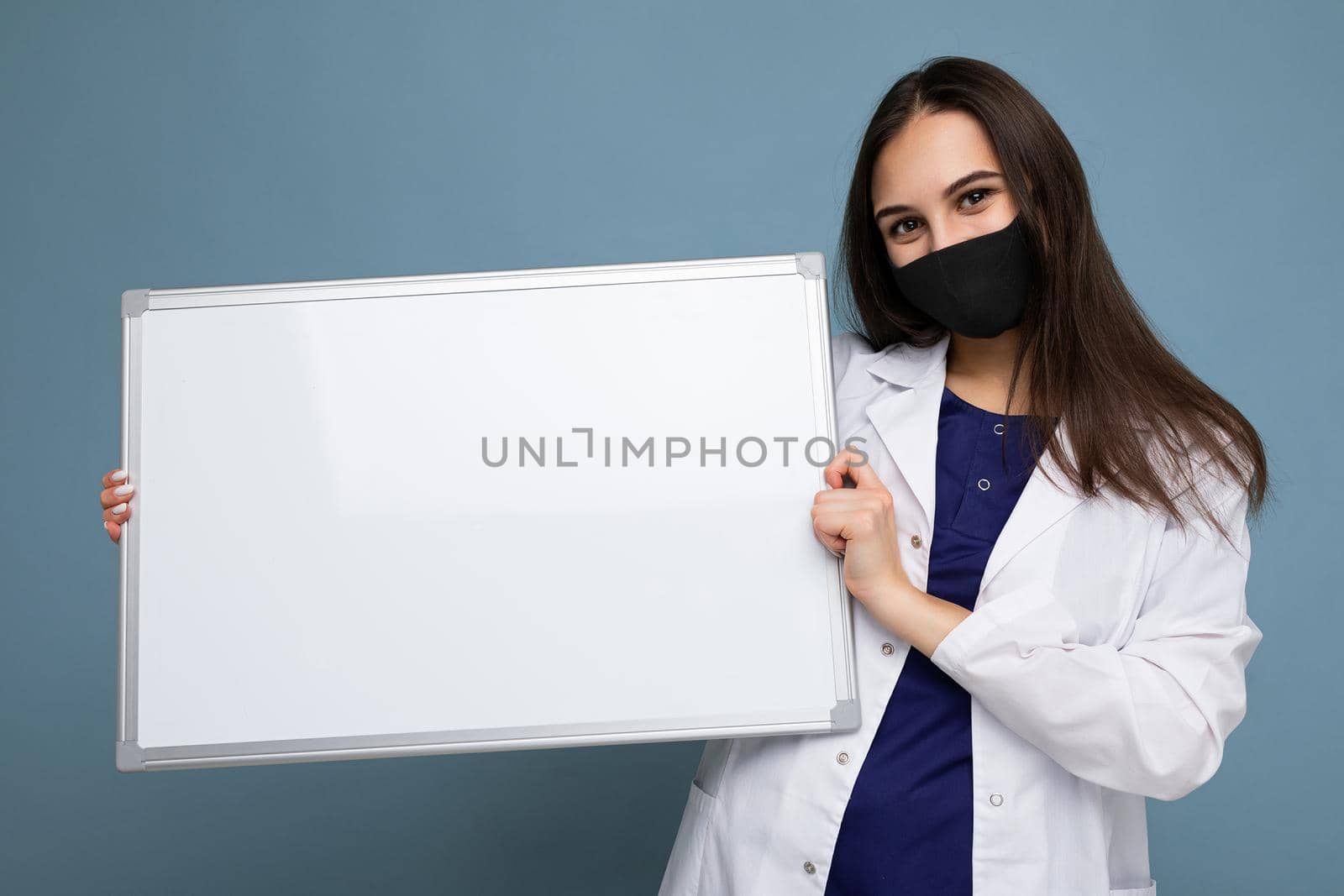 Woman doctor wearing a white medical coat and a mask holding blank board with copy space for text isolated on background. Coronavirus concept by TRMK