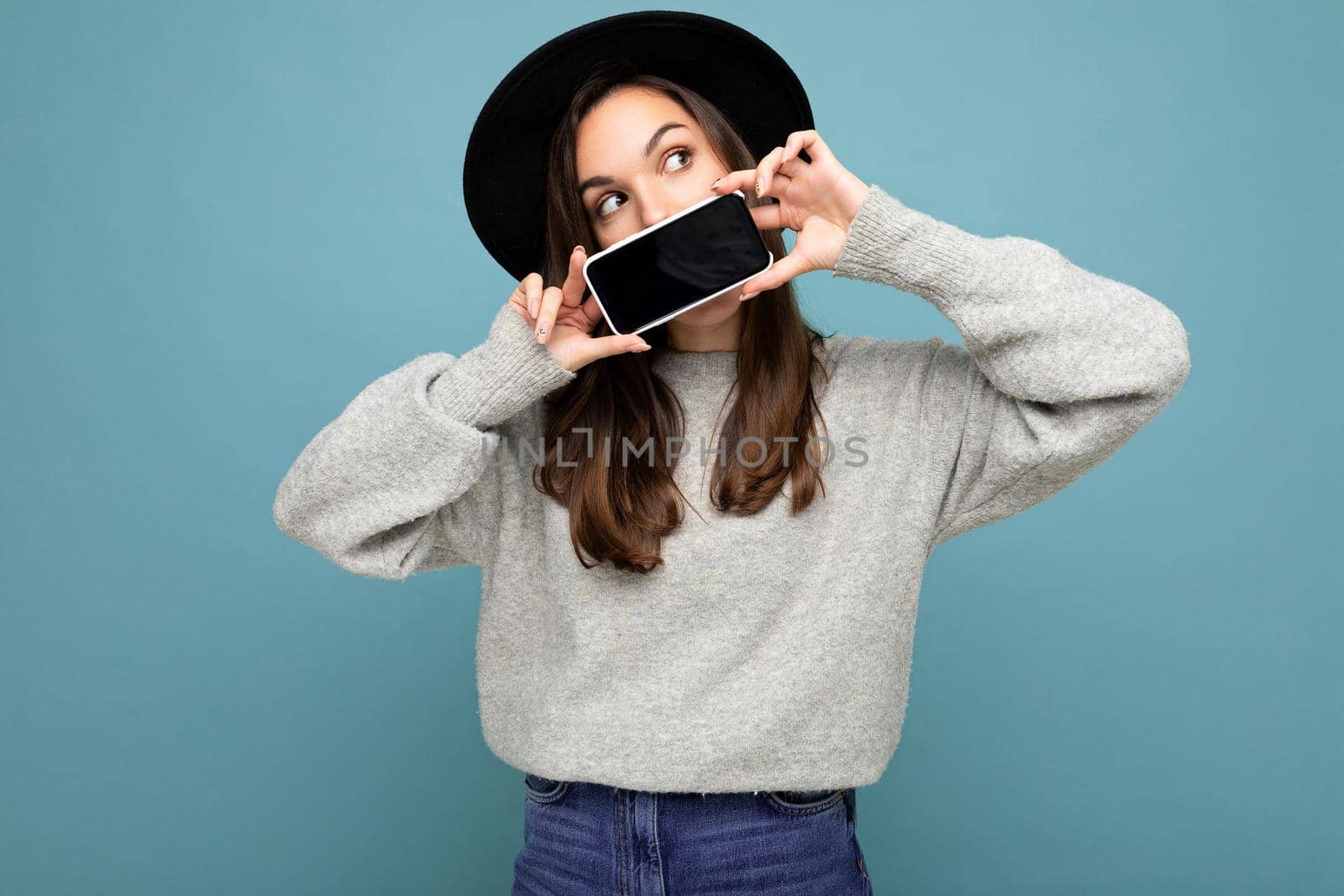Photo of Beautiful positive woman person wearing black hat and grey sweater holding mobilephone showing smartphone isolated on background lookingto the side.Mock up, cutout, free space