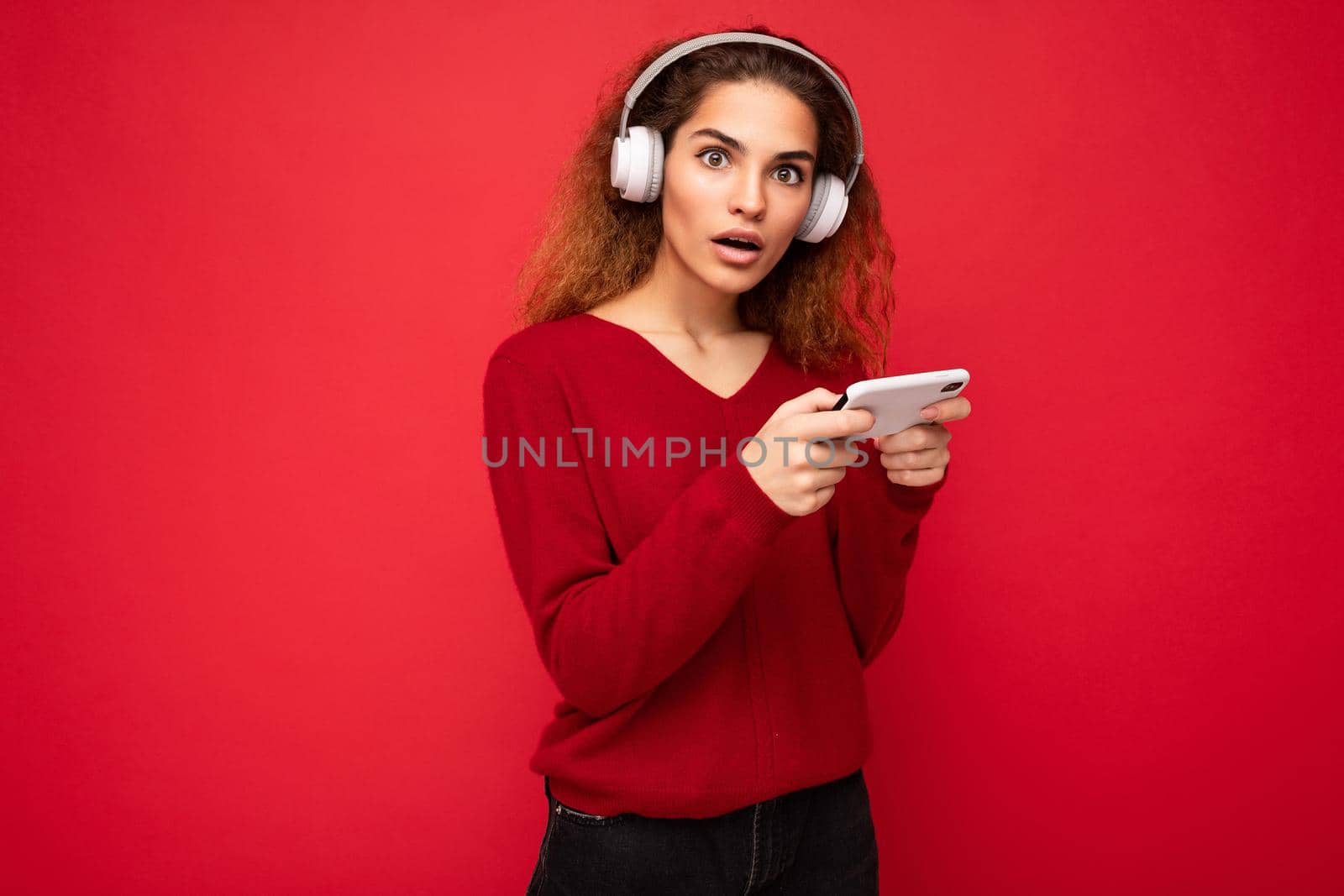 Pretty amazed with open mouth young brunette curly woman wearing dark red sweater isolated on red backdrop wearing white headphones listening to music and using smartphone looking at camera by TRMK