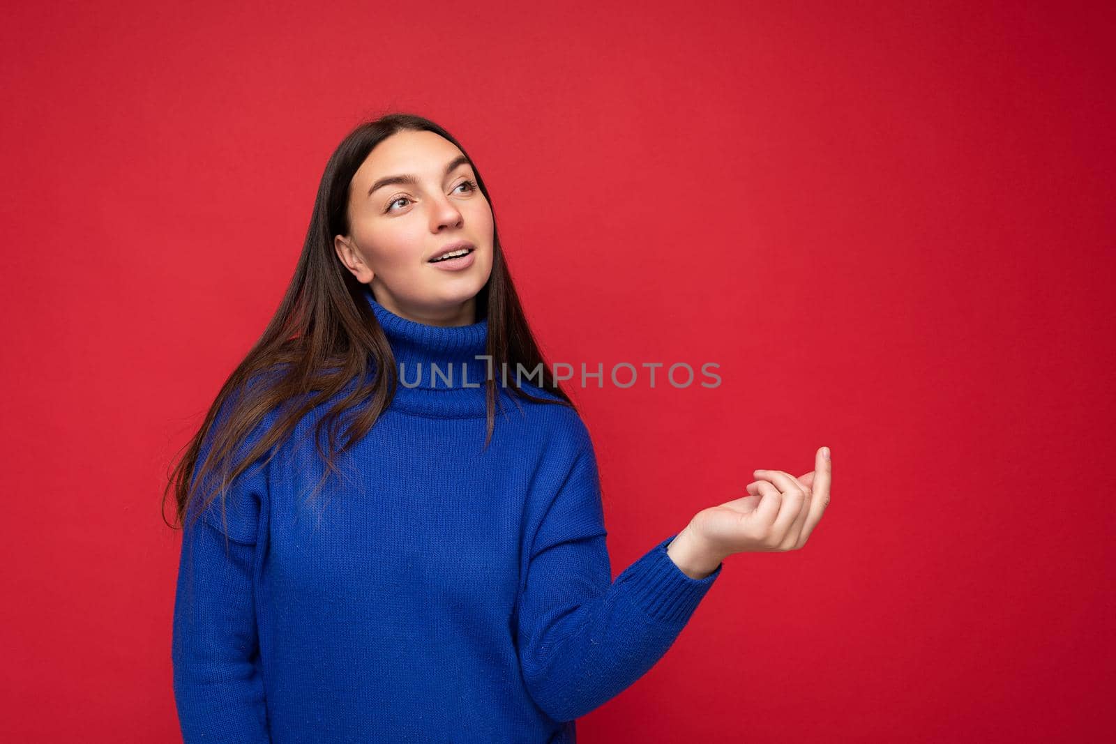 Portrait of positive young attractive brunette woman with sincere emotions wearing trendy bright blue pullover isolated over red background with free space.