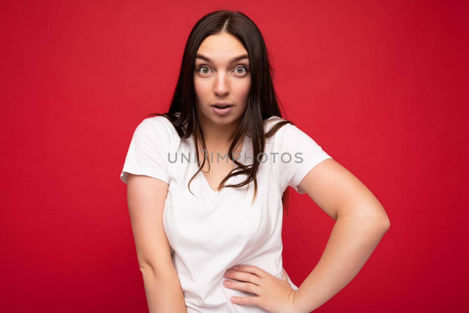 Photo of surprised young beautiful brunet lady with sincere emotions wearing casual white t-shirt for mockup isolated over red background with copy space.