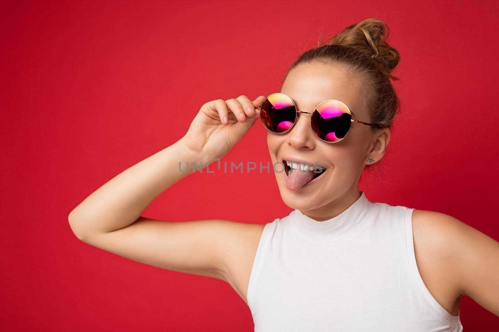 Photo shot of beautiful happy young blonde woman wearing casual clothes and stylish sunglasses isolated over colorful background looking at camera and showing tongue.
