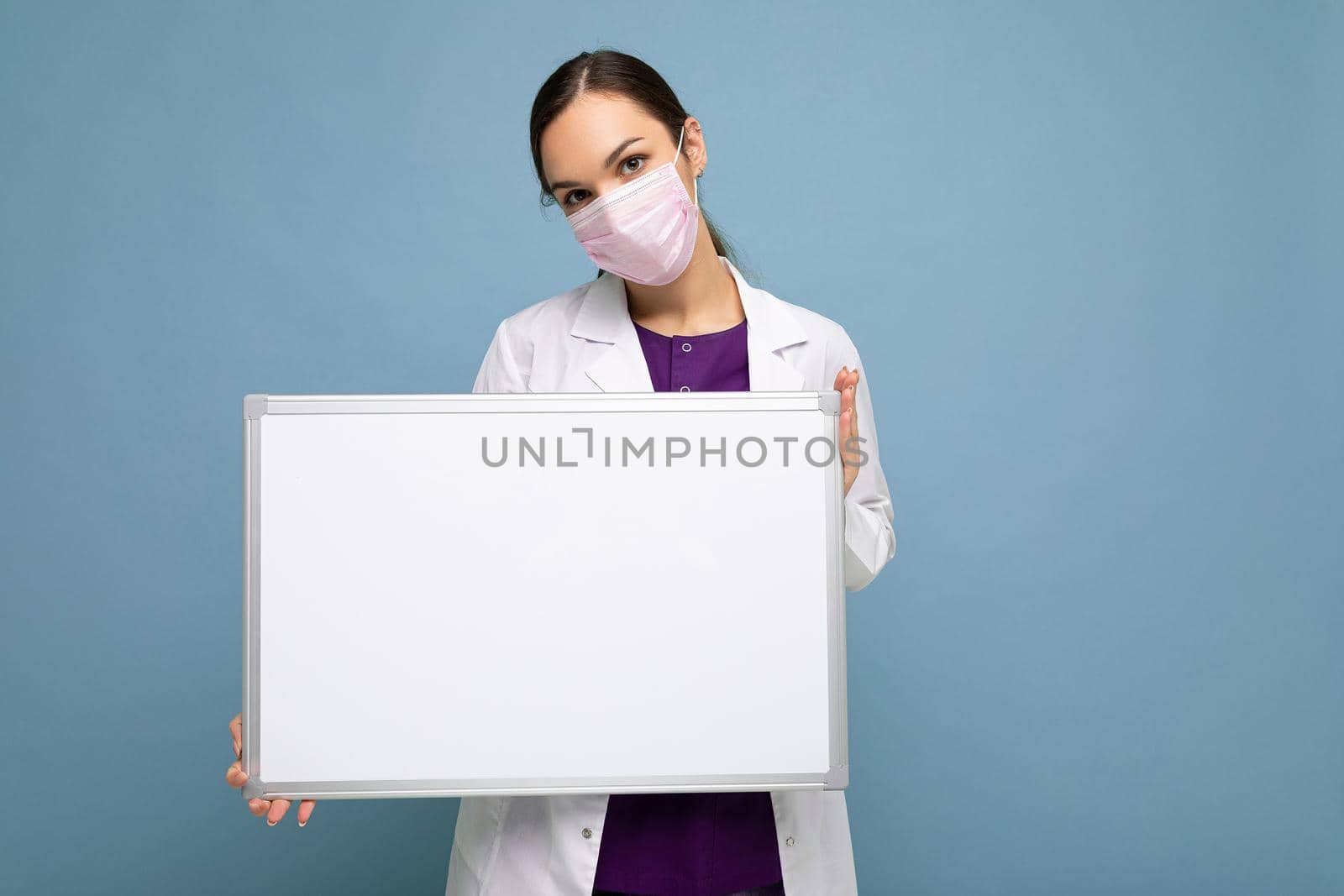 Young attractive nurse in protective face mask and white medical coat holding an empty magnetic board isolated on blue background by TRMK