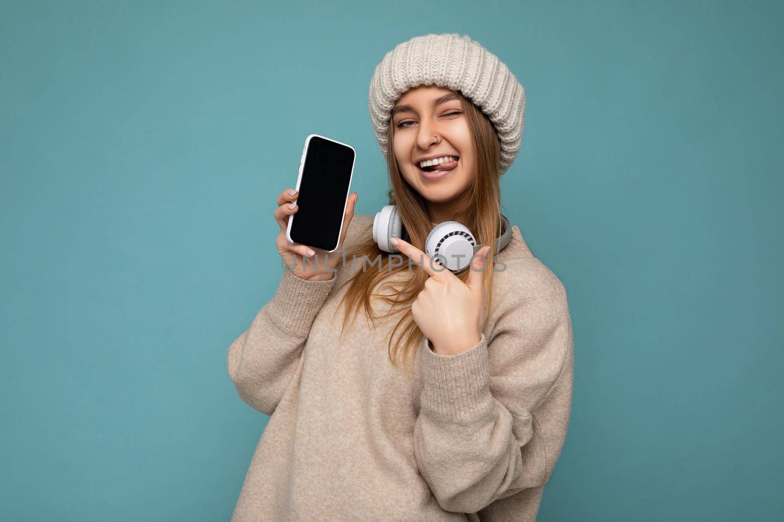 Photo of sexy attractive positive joyful young dark blonde woman wearing beige stylish sweater and knitted beige winter hat isolated on blue background wearing white bluetooth wireless earphones and listening to cool music and showing mobile phone with empty display for mockup looking at camera and pointing at device.