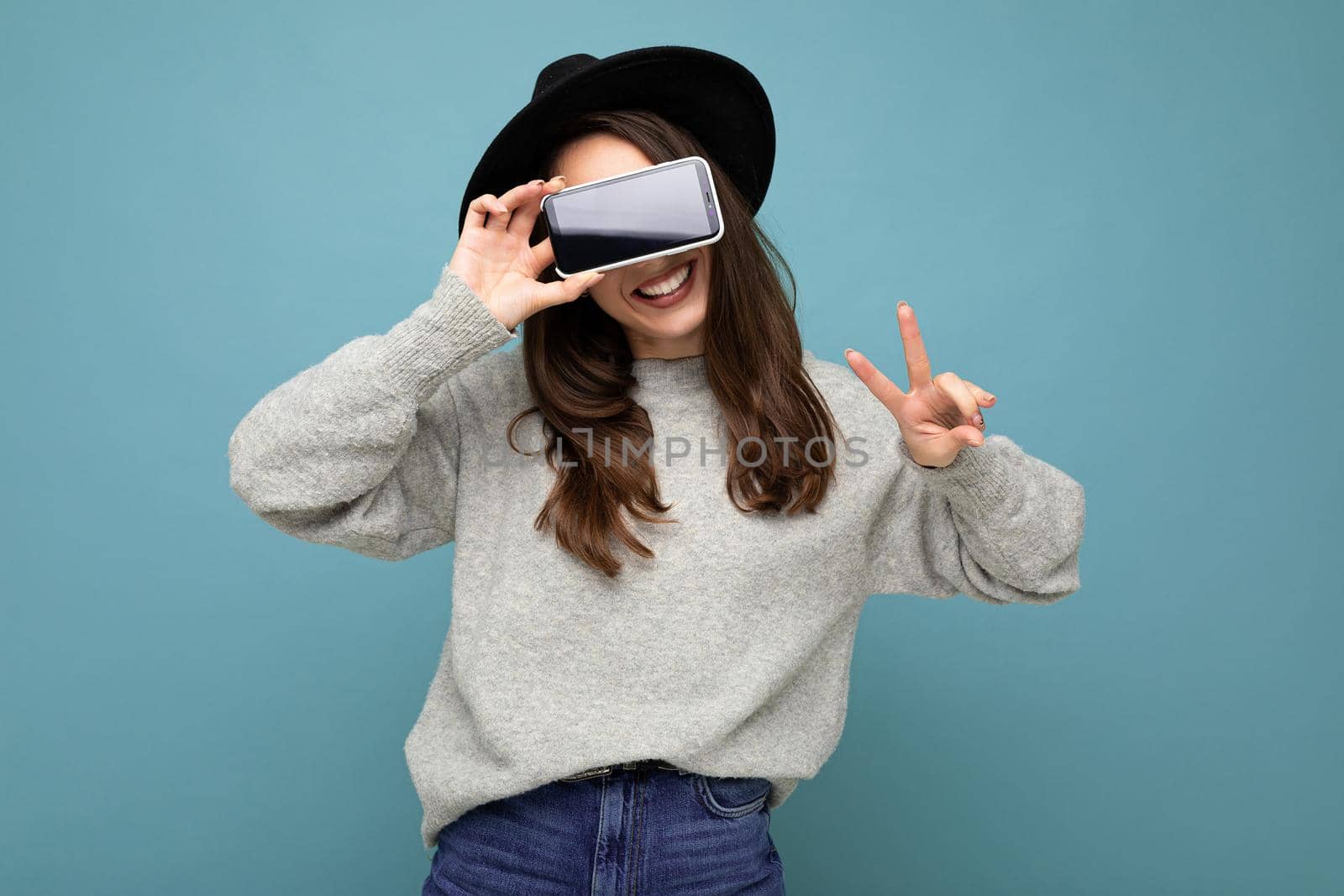 Pretty positive young female person wearing black hat and grey sweater holding phone showing smartphone isolated on background showing peace gesture by TRMK