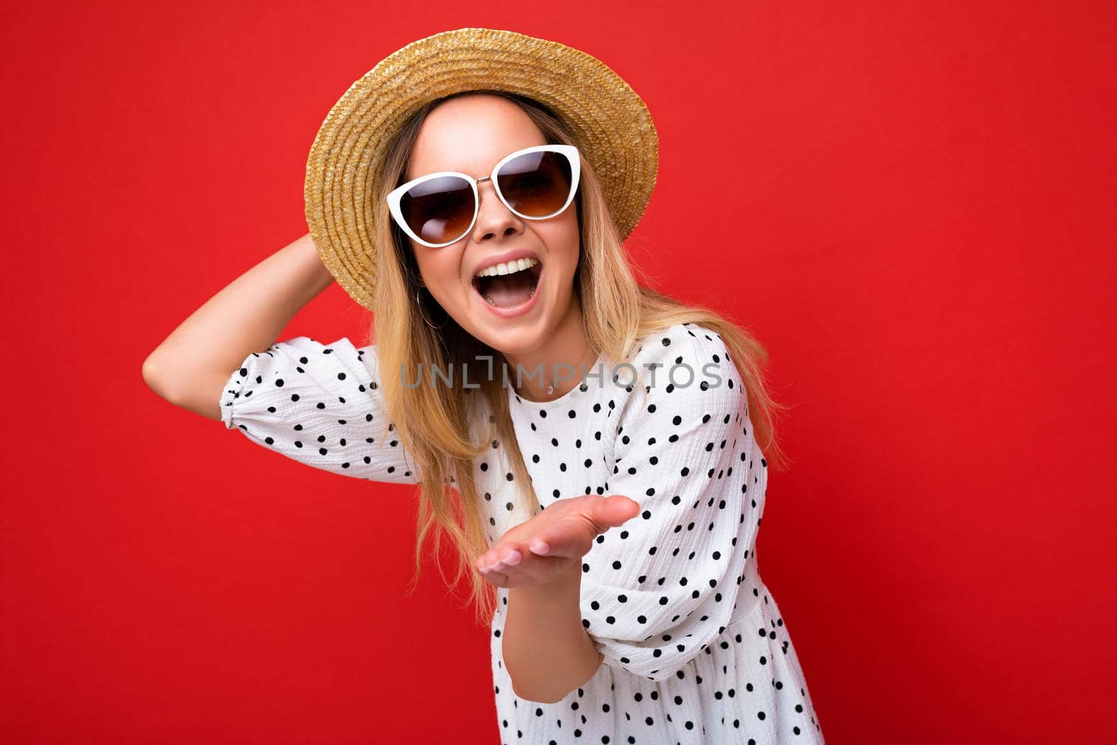 Shot of pretty positive smiling young blonde woman wearing summer dress straw hat and stylish glasses isolated over red background wall looking at camera and giving kiss by TRMK