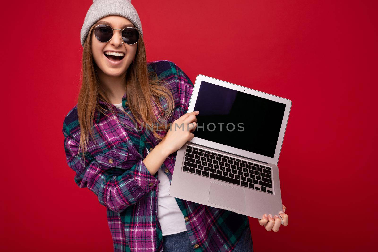Photo shot of amazing funny happy beautiful smiling dark blond young woman holding computer laptop with empty monitor screen with mock up and copy space wearing sun glasses hat and colourful shirt looking at camera isolated over red wall background.