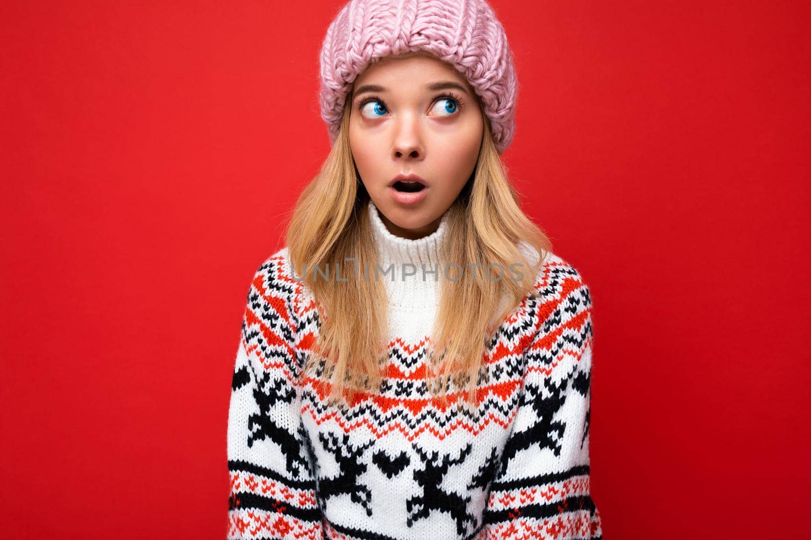 Shot of beautiful astonished young blonde woman isolated over red background wall wearing winter sweater and pink hat looking up to the side by TRMK