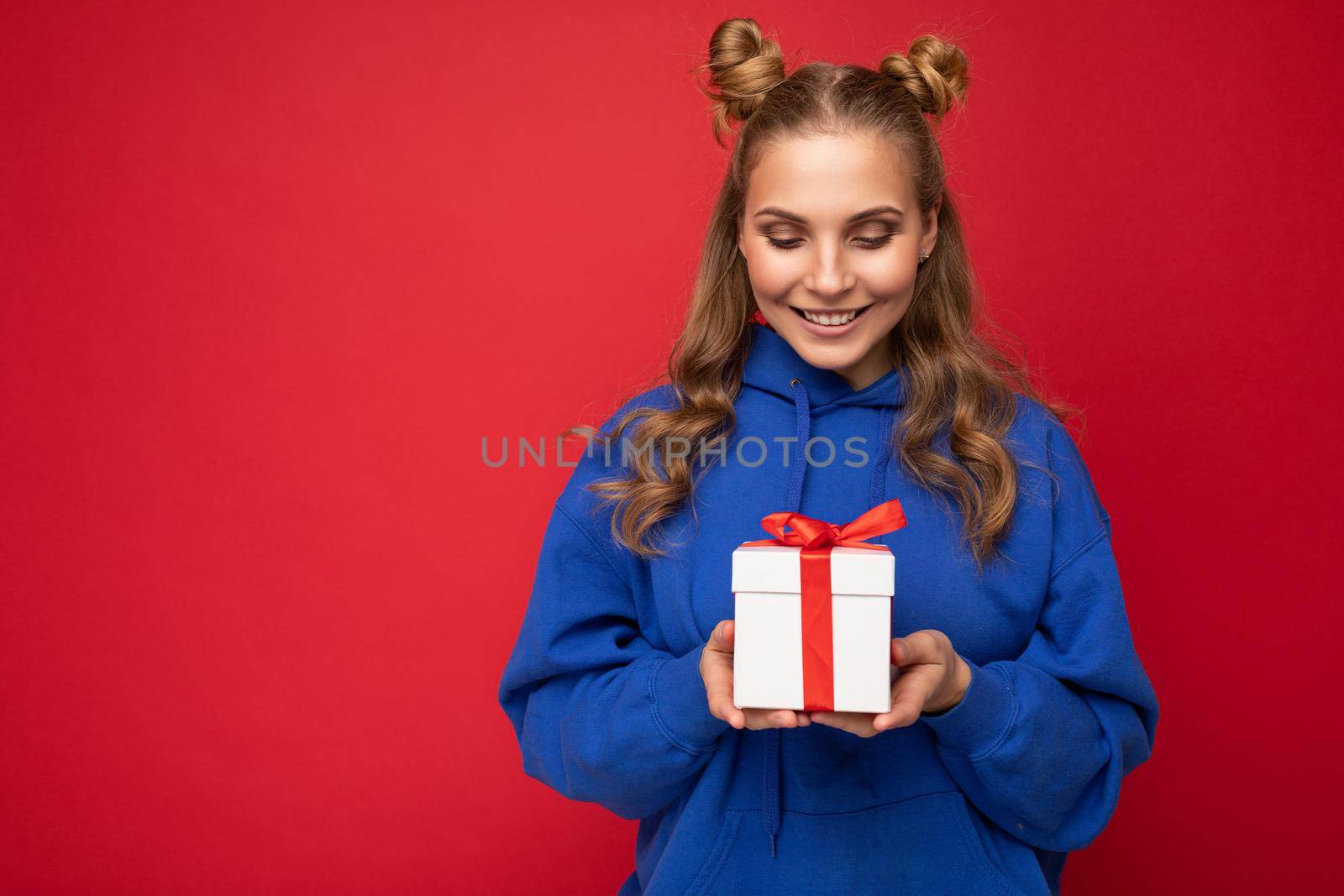 Shot of attractive happy smiling young blonde woman isolated over red background wall wearing blue trendy hoodie holding gift box and looking at white box with red ribbon by TRMK