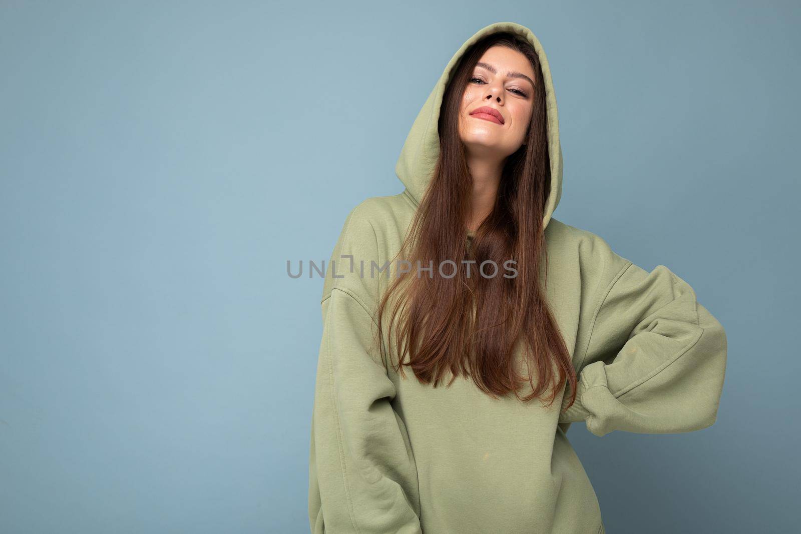 Young beautiful woman looking at camera. Trendy girl in casual hipster hoodie clothes. Positive female shows facial emotions. Funny model isolated on blue background with free space for text by TRMK