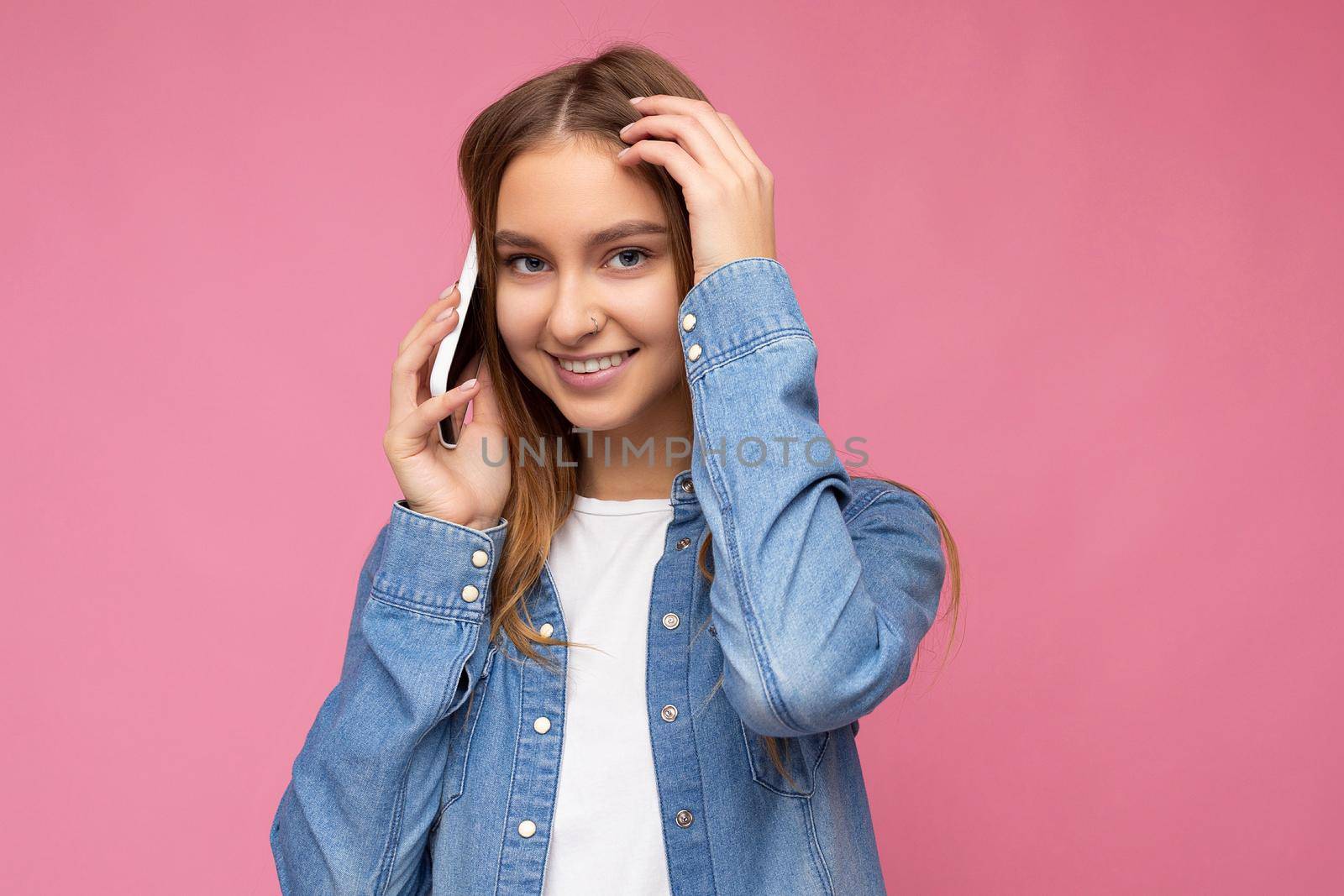 Photo of beautiful smiling happy young blonde woman wearing casual blue jean shirt isolated over pink background holding in hand and communicating on mobile phone looking at camera.