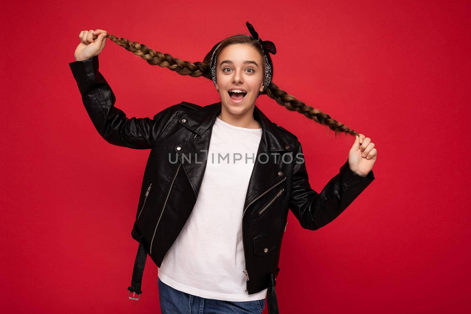 Photo shot of charming positive amazing brunet little female teenager with pigtails wearing stylish black leather jacket and white t-shirt for mockup standing isolated over red background wall looking at camera.
