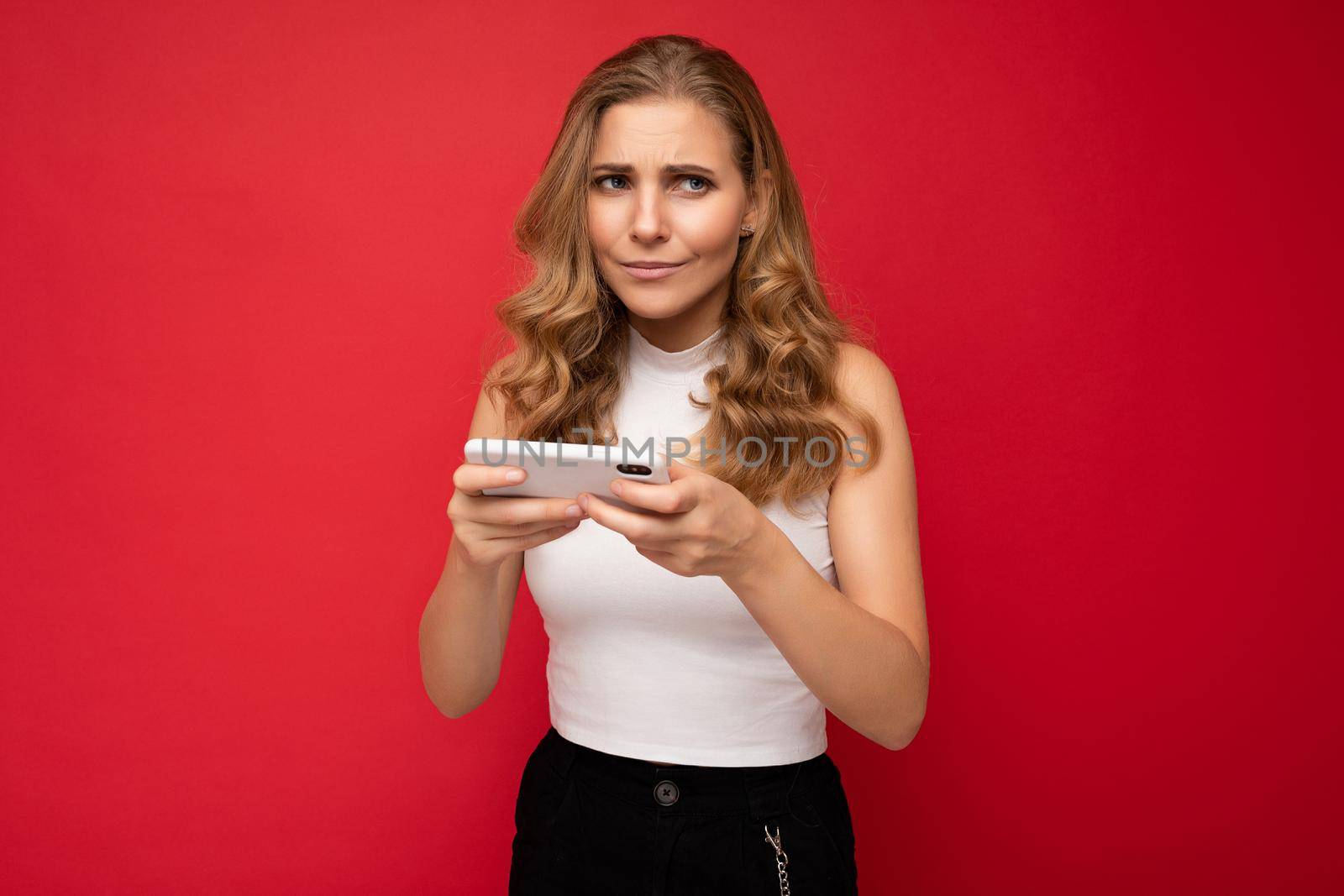 Photo of concentrated beautiful young blonde woman wearing white t-shirt isolated on red background using smartphone and playing phone games looking to the side.