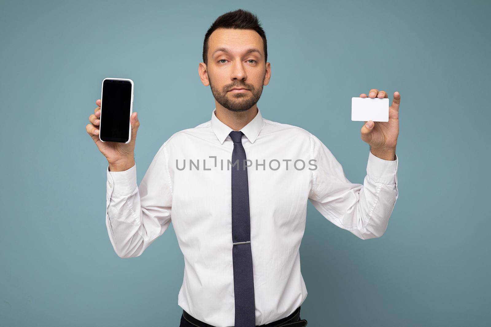 Photo of handsome good looking brunet man with beard wearing casual white shirt and tie isolated on blue background with empty space holding in hand and showing mobile phone with empty screen for mockup and credit card looking at camera by TRMK