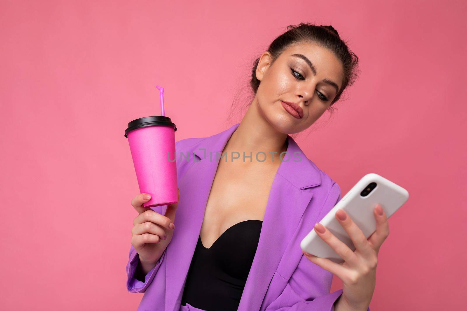 Shot of dissatisfied sad upset caucasian brunette woman holds take away coffee dressed in casual clothes isolated over pink wall uses smartphone. Negative concept by TRMK