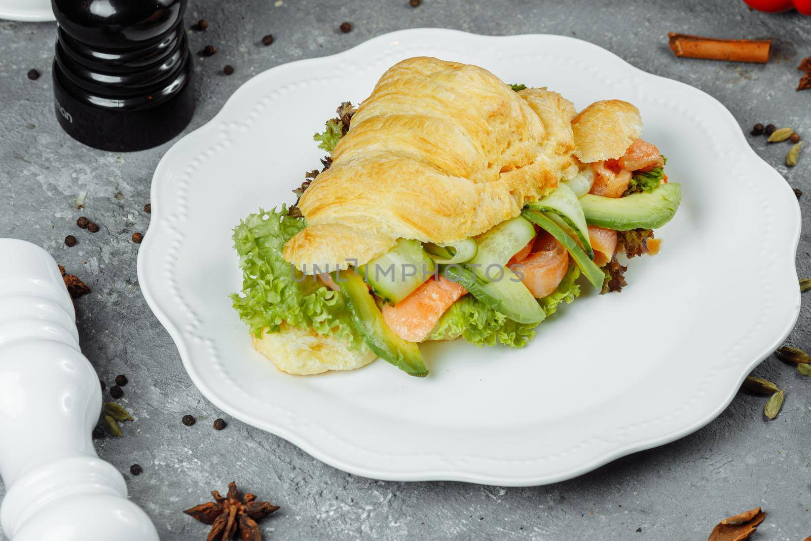 Croissant sandwich with red fish, avocado, fresh vegetables and arugula on black shale board over black stone background. Healthy food concept by UcheaD