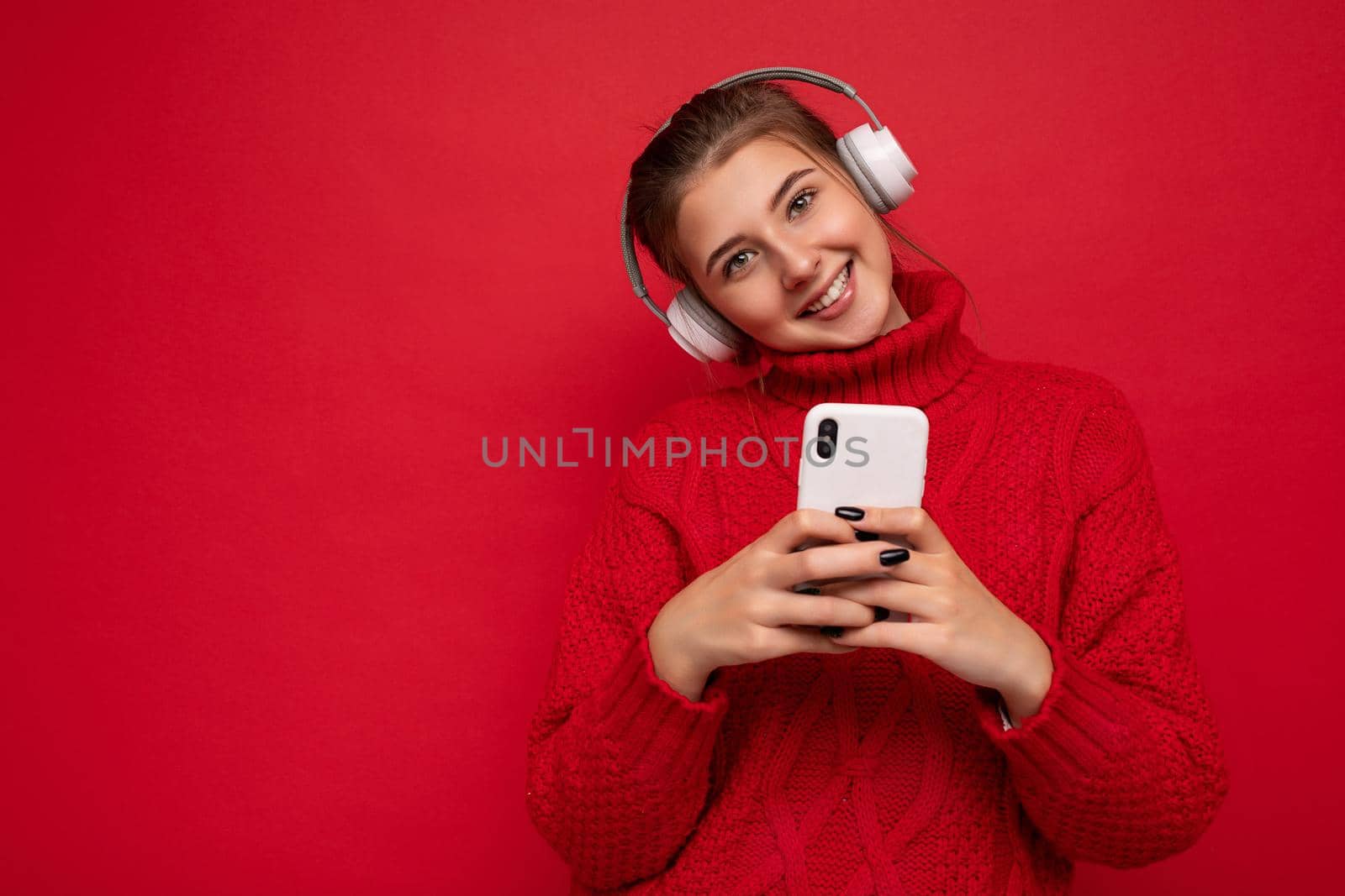 Photo of beautiful joyful smiling young woman wearing stylish casual clothes isolated over background wall holding and using mobile phone wearing white bluetooth headphones listening to music and having fun looking at camera by TRMK