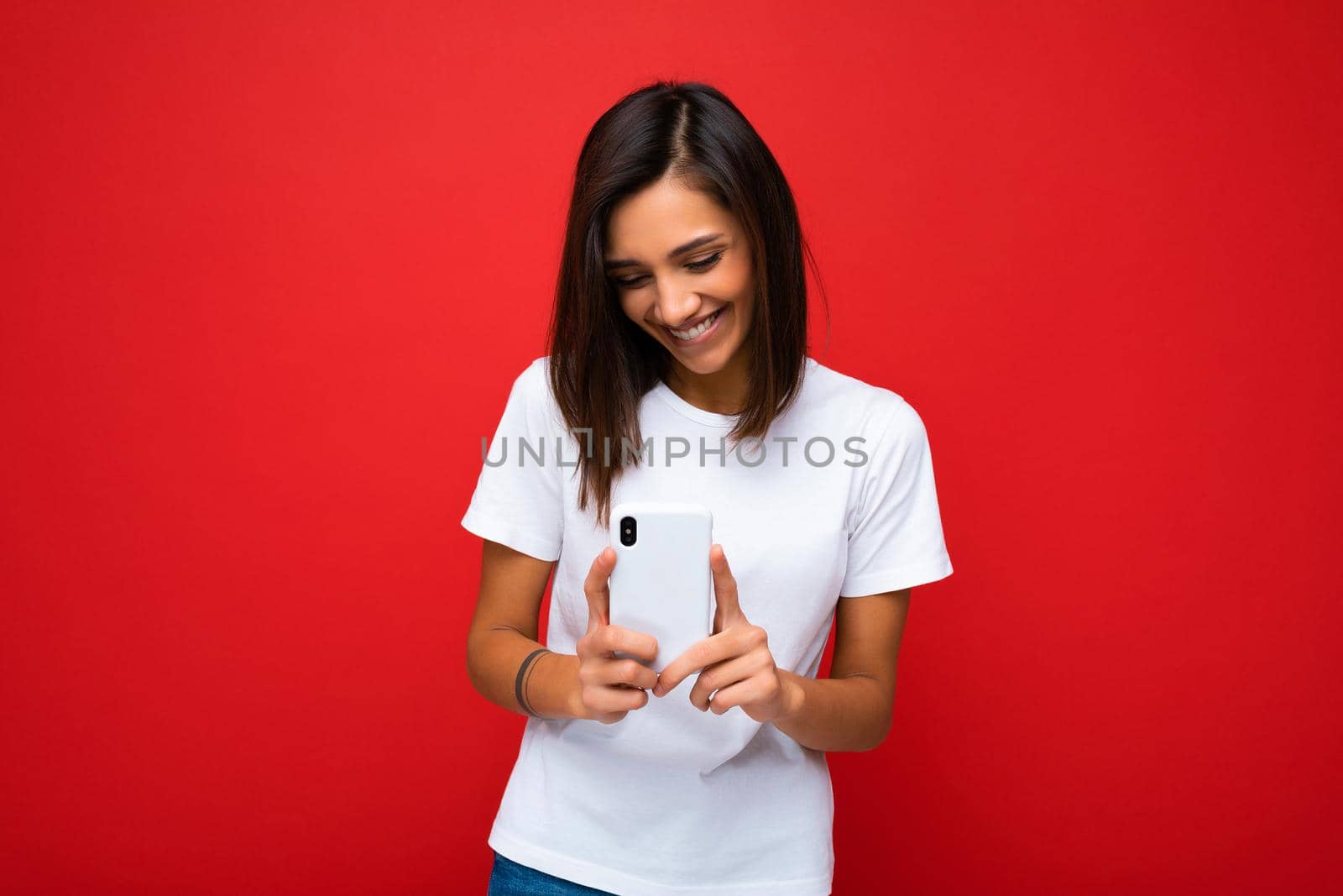 Photo shot of attractive positive good looking young woman wearing casual stylish outfit poising isolated on background with empty space holding in hand and using mobile phone messaging sms looking at smartphone display screen.