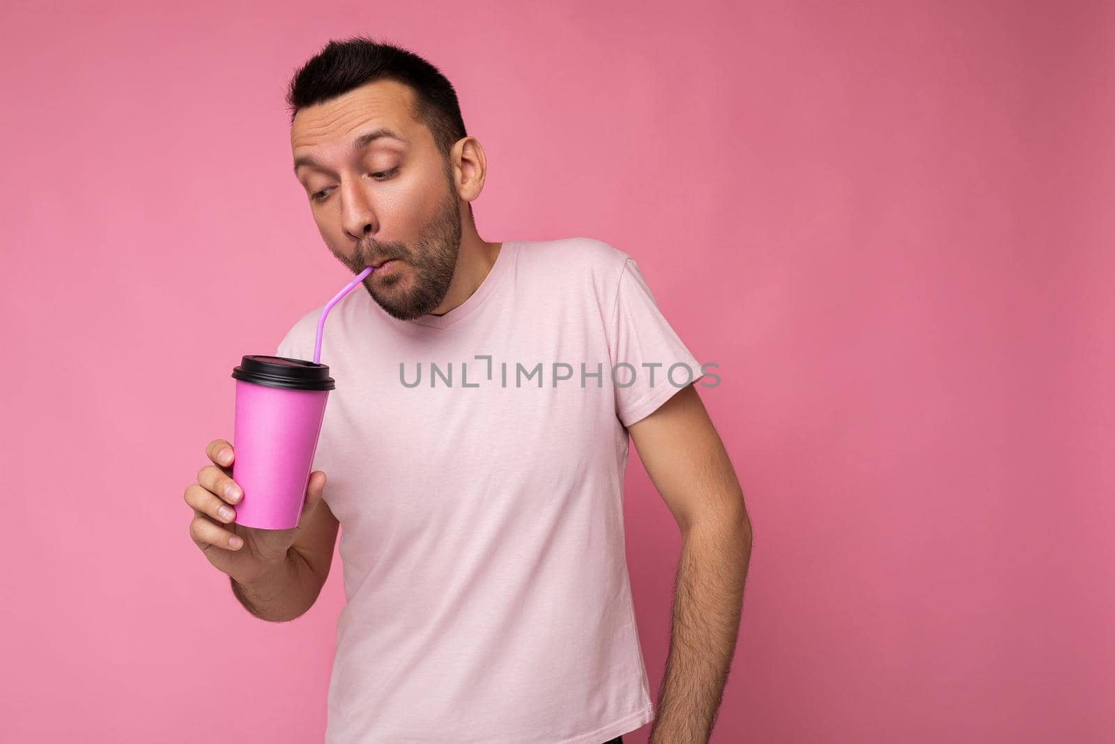 Closeup photo of Handsome funny joyful young brunette unshaven male person with beard wearing white t-shirt isolated over pink backgroung wall holding paper cup drinking and enjoying. empty space