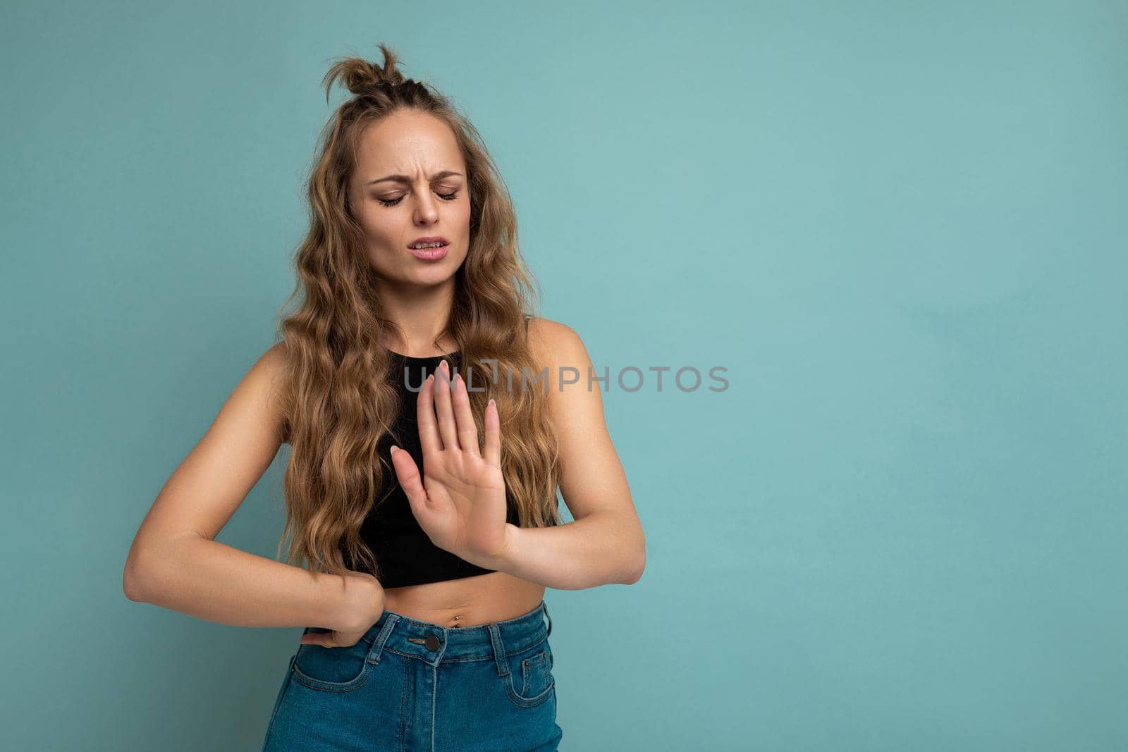 Young dissatisfied attractive winsome blonde curly woman with sincere emotions wearing trendy black top isolated over blue background with free space and showing stop sign. Negative concept.