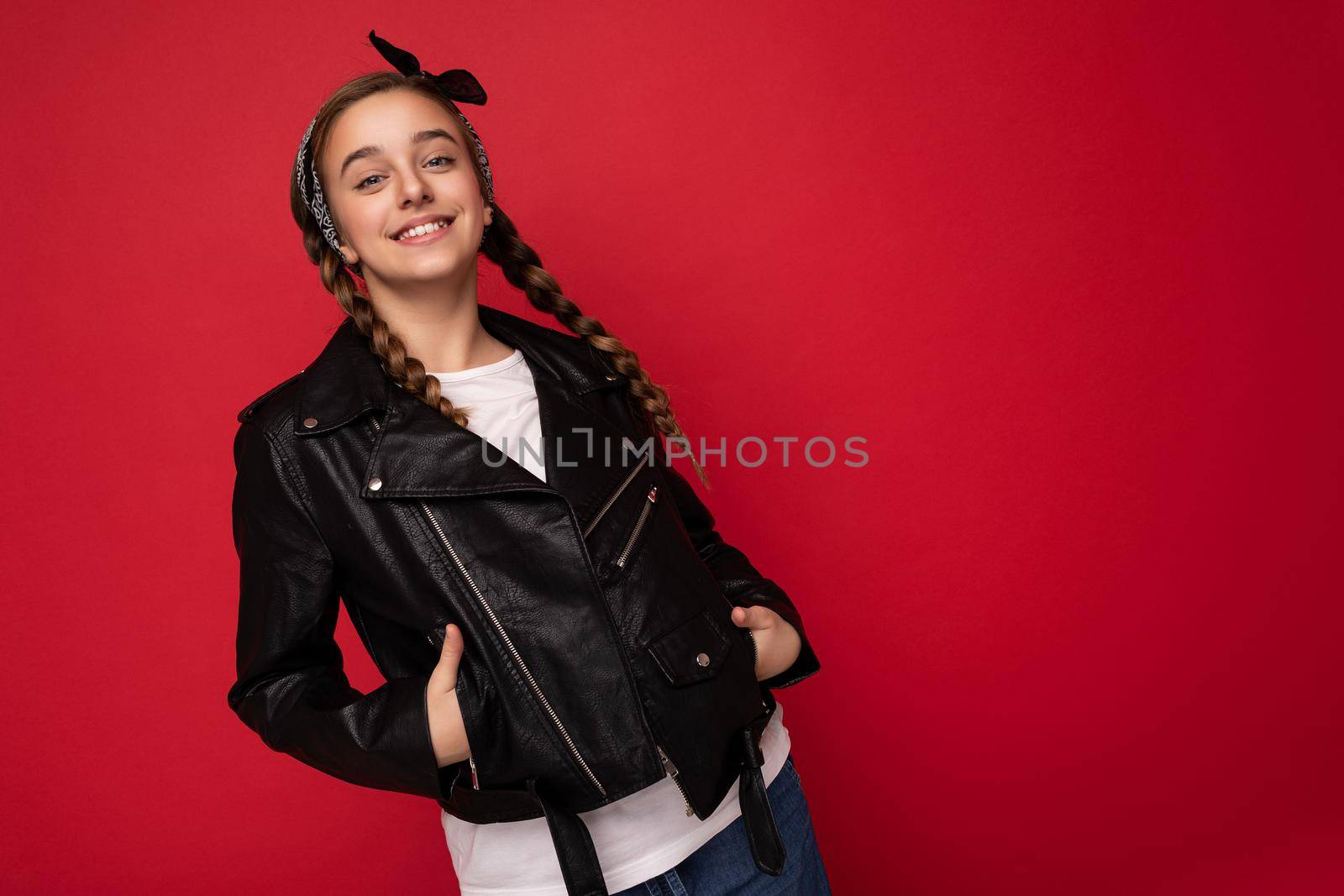 Shot of attractive cool positive smiling brunette little female teenager with pigtails wearing stylish black leather jacket and white t-shirt isolated on red background looking at camera by TRMK