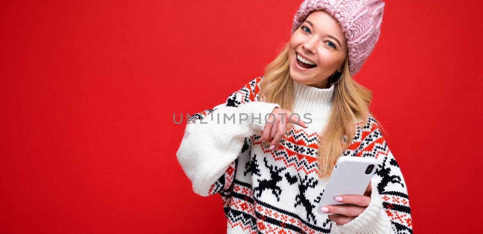 Panoramic Photo of beautiful smiling young blonde woman wearing warm knitted hat and winter warm sweater standing isolated over red background communicating online via smartphone looking at camera by TRMK