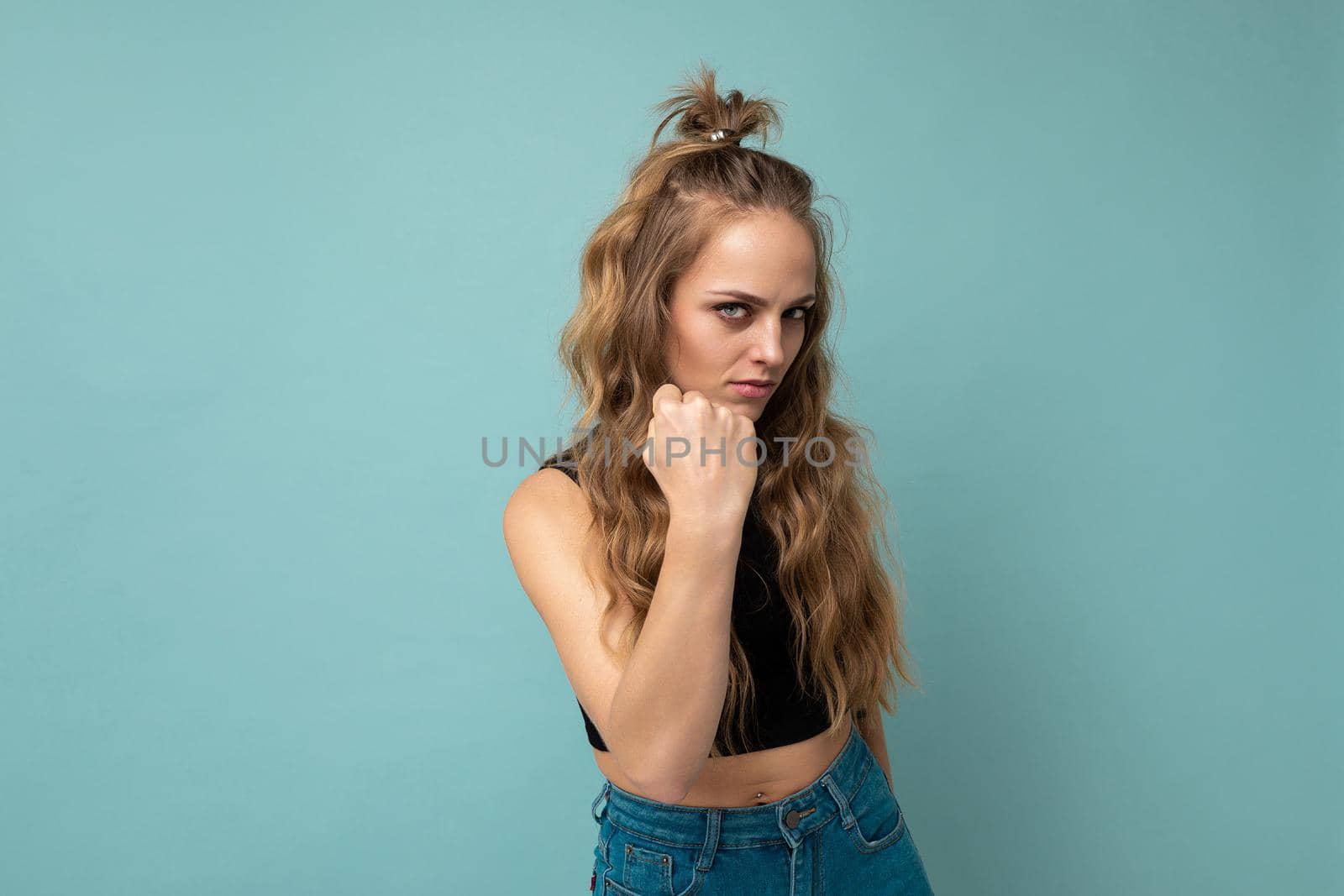 Young dissatisfied attractive winsome blonde curly woman with sincere emotions wearing trendy black top isolated over blue background with free space and showing fist. Negative concept.