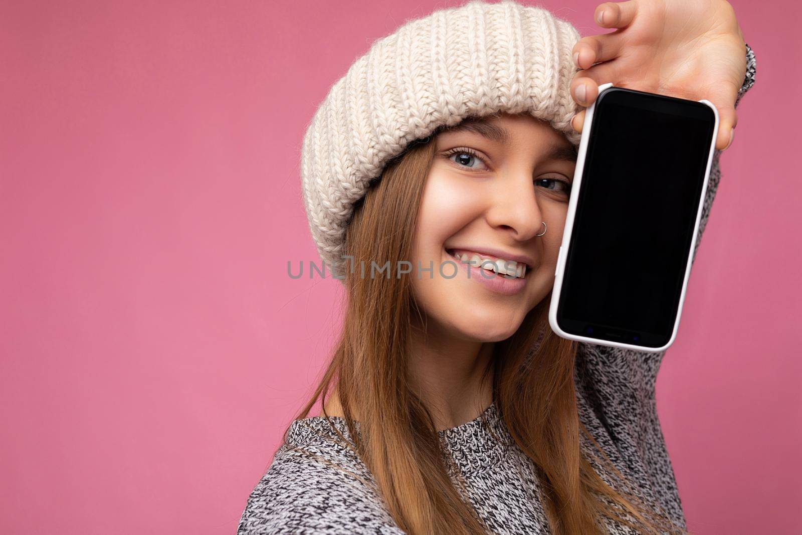 Closeup photo of Beautiful smiling delightful young blonde woman wearing casual grey sweater and beige hat isolated over pink background holding in hand and showing mobile phone with empty display for mockup looking at camera.