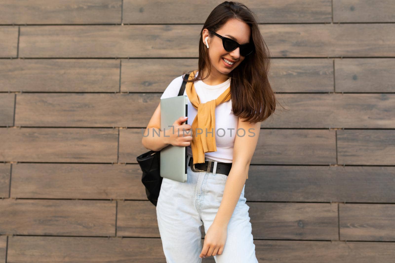 Fascinating beautiful smiling charming young brunet girl looking down holding computer laptop and black sunglasses in white t-shirt and light blue jeans in the street near brown wall by TRMK