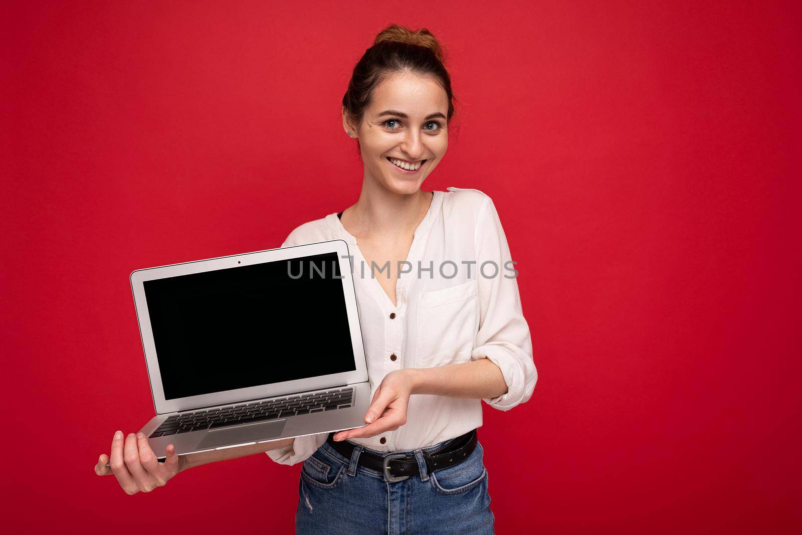 portrait of Beautiful smiling happy young woman holding computer laptop looking at camera having fun wearing casual smart clothes isolated over red wall background. copy space