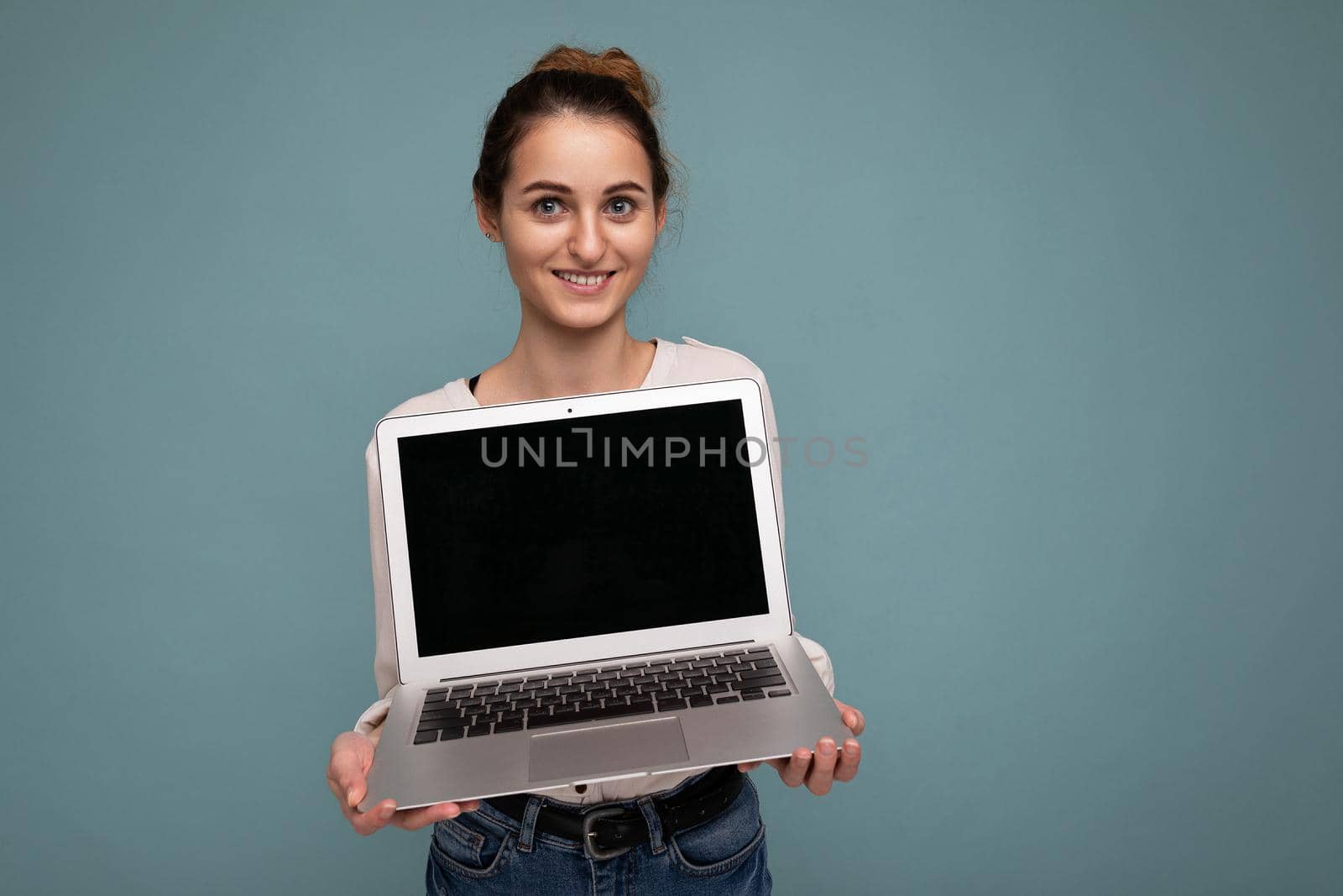 Beautiful smiling young woman holding netbook computer looking at camera wearing white shirt isolated on blue background. mock up