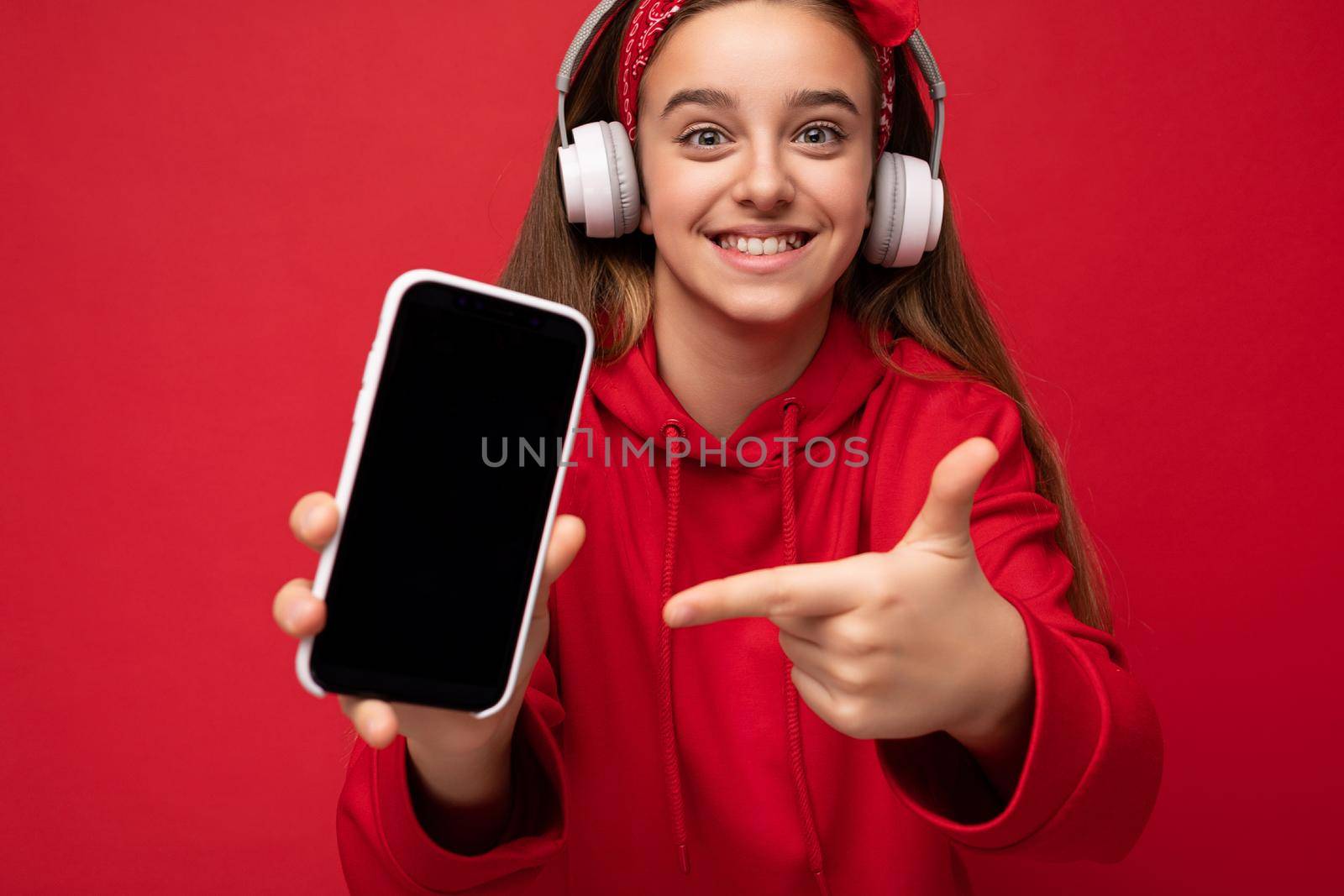 Closeup Photo of positive smiling cute brunette girl wearing red hoodie isolated on red background holding and showing smartphone with empty display for cutout wearing white wireless headphones listening to funny music looking at camera and pointing finger at gadjet.