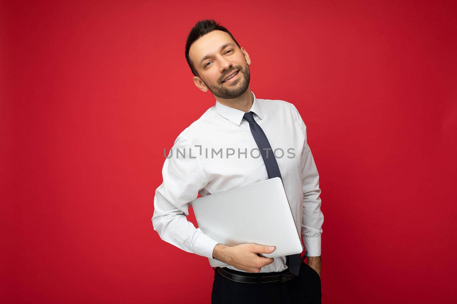 Handsome smiling brunet man holding laptop computer looking at camera in white shirt and tie on isolated red background.