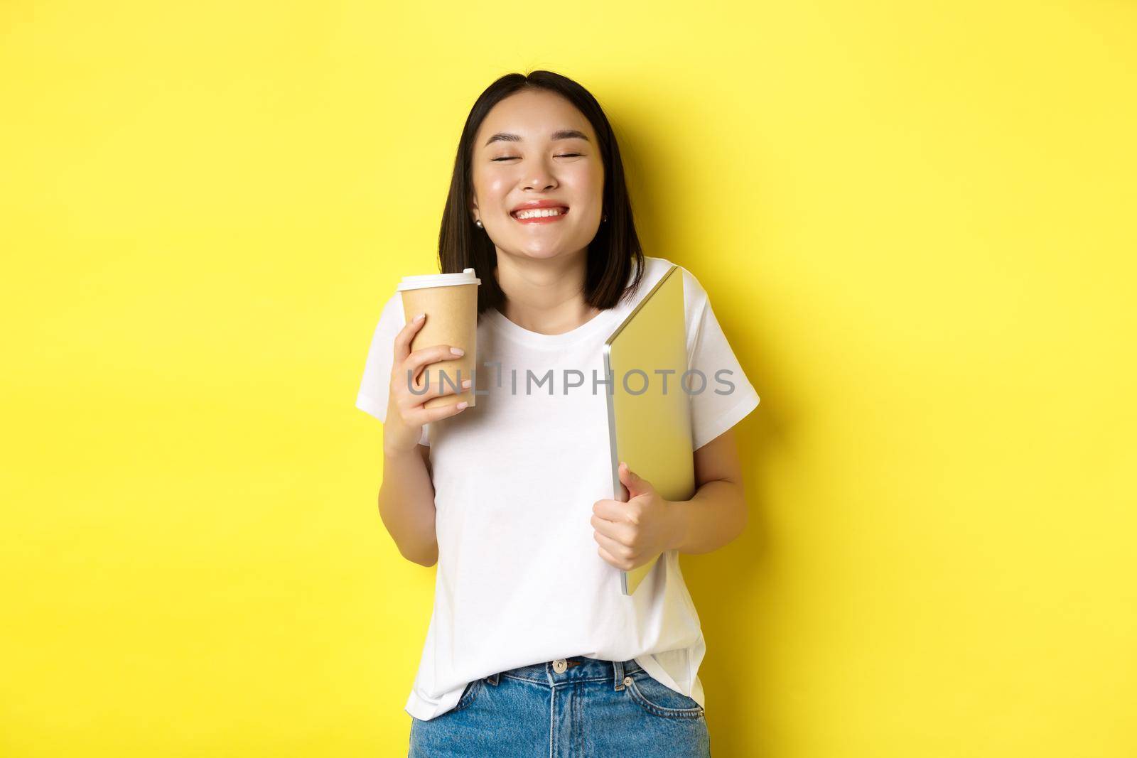 Happy young woman enjoying coffee from cafe and smiling, holding cup and laptop, standing over yellow background.