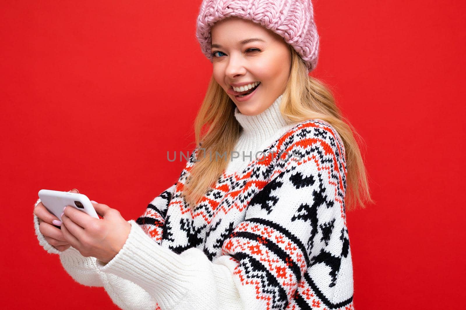 closeup Photo of winking beautiful smiling young blonde woman wearing warm knitted hat and winter warm sweater standing isolated over red background playing games via smartphone looking at camera.